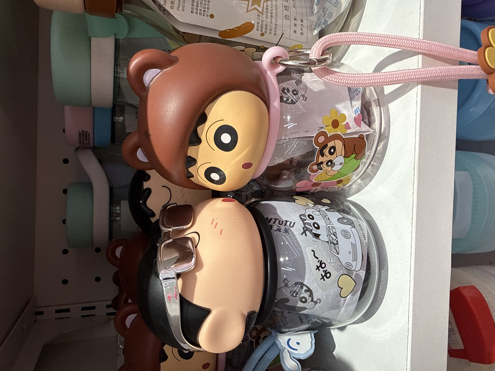 Two cartoon character figurines, possibly part of a merchandise display in a store, are positioned on a white shelf. The figurine on the left, wearing sunglasses and a black headpiece, has a clear plastic body filled with paper. A blue fabric rabbit charm is attached to its side. The figurine on the right wears a brown hood with bear ears and has a pink scarf. Its clear plastic body is also filled with paper, and it has a pink rope with a metal ring and a bear charm hanging from its neck. Behind the figurines, other similar items and shelves are visible, including what appear to be plastic bottles and containers with colorful lids. The environment suggests a retail setting, possibly in the city of Xiaoshan District, China, given the presence of Chinese text on the packaging.