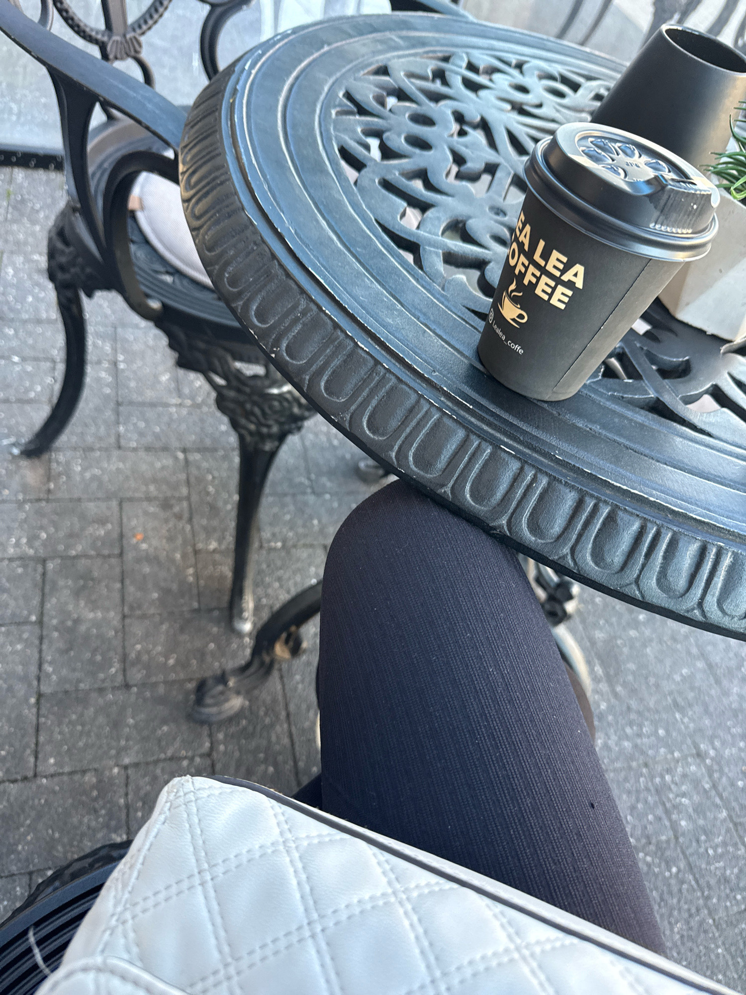 A close-up, high-angle outdoor shot shows a black disposable coffee cup with a black lid on a black ornate metal cafe table. The text on the cup reads "LEA LEA COFFEE" in gold lettering. The cup is positioned to the right of center on the table, angled slightly towards the viewer. To the left of the cup, a portion of a person's legs, clad in dark leggings, is visible, along with the corner of a white quilted handbag. The table appears to be set up on a paved sidewalk or plaza, with cobblestone-like paving stones visible in the background. The overall impression is of a casual cafe setting in an urban environment. The location is identified as a cafe in Chișinău, Moldova, as per the prompt's instructions.