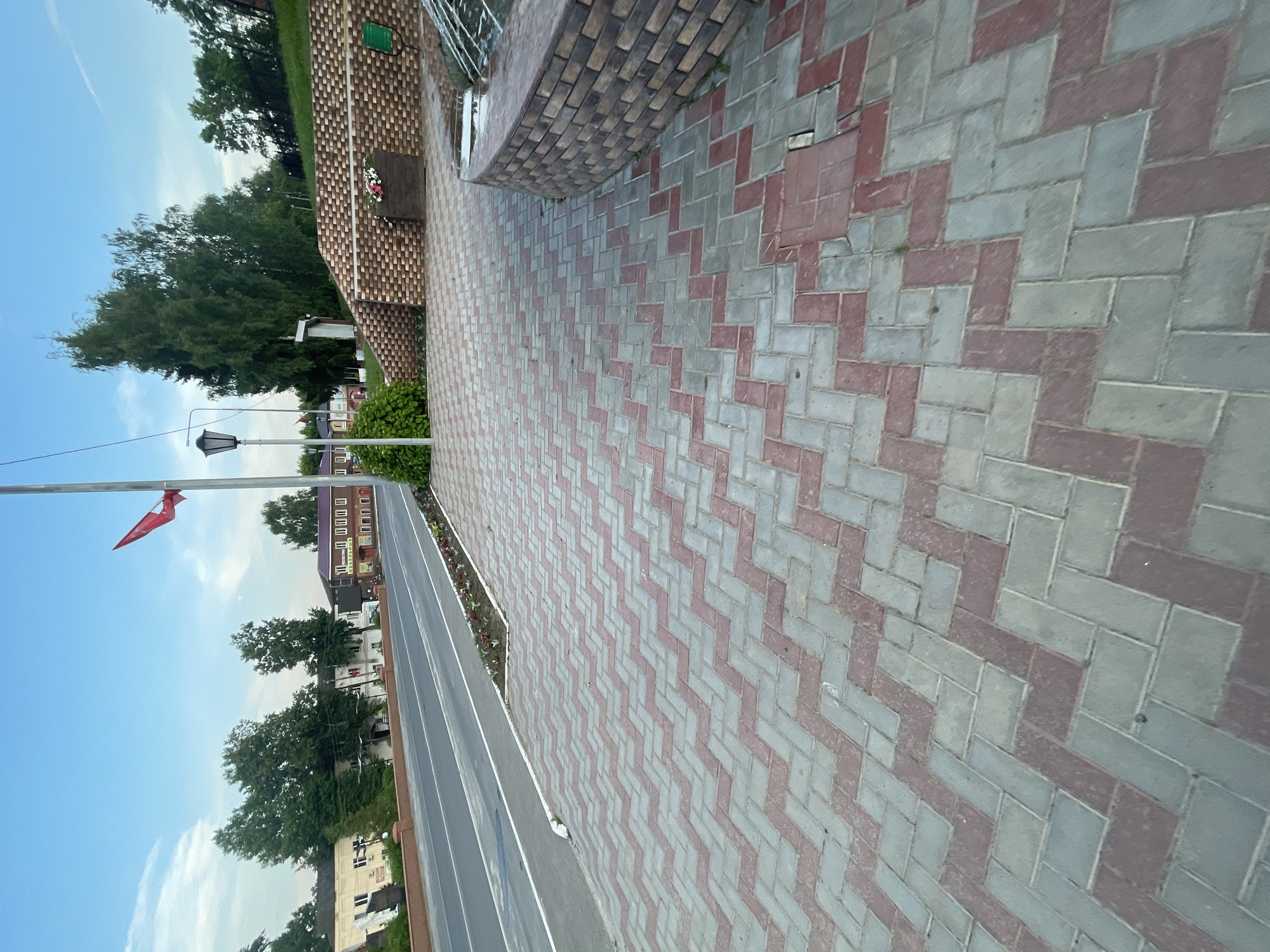 The image depicts a street scene in the town of Krasnye Baki, Russia, under a clear blue sky with scattered clouds. The foreground is dominated by a red and gray patterned interlocking brick walkway. To the left, a paved road runs parallel to the walkway, with a white line marking its edge. Beyond the road, there are several buildings, including a multi-story building with a light-colored facade and a red-roofed building with windows and signage. A red flag is visible on a pole in the distance, and several trees are scattered throughout the scene. To the right, a brick wall curves and forms part of what appears to be a water feature or fountain, with water visible. A dark-colored car is partially visible in the lower right corner of the image. The overall impression is one of a public area in a town, likely a street or a park bordering a road.