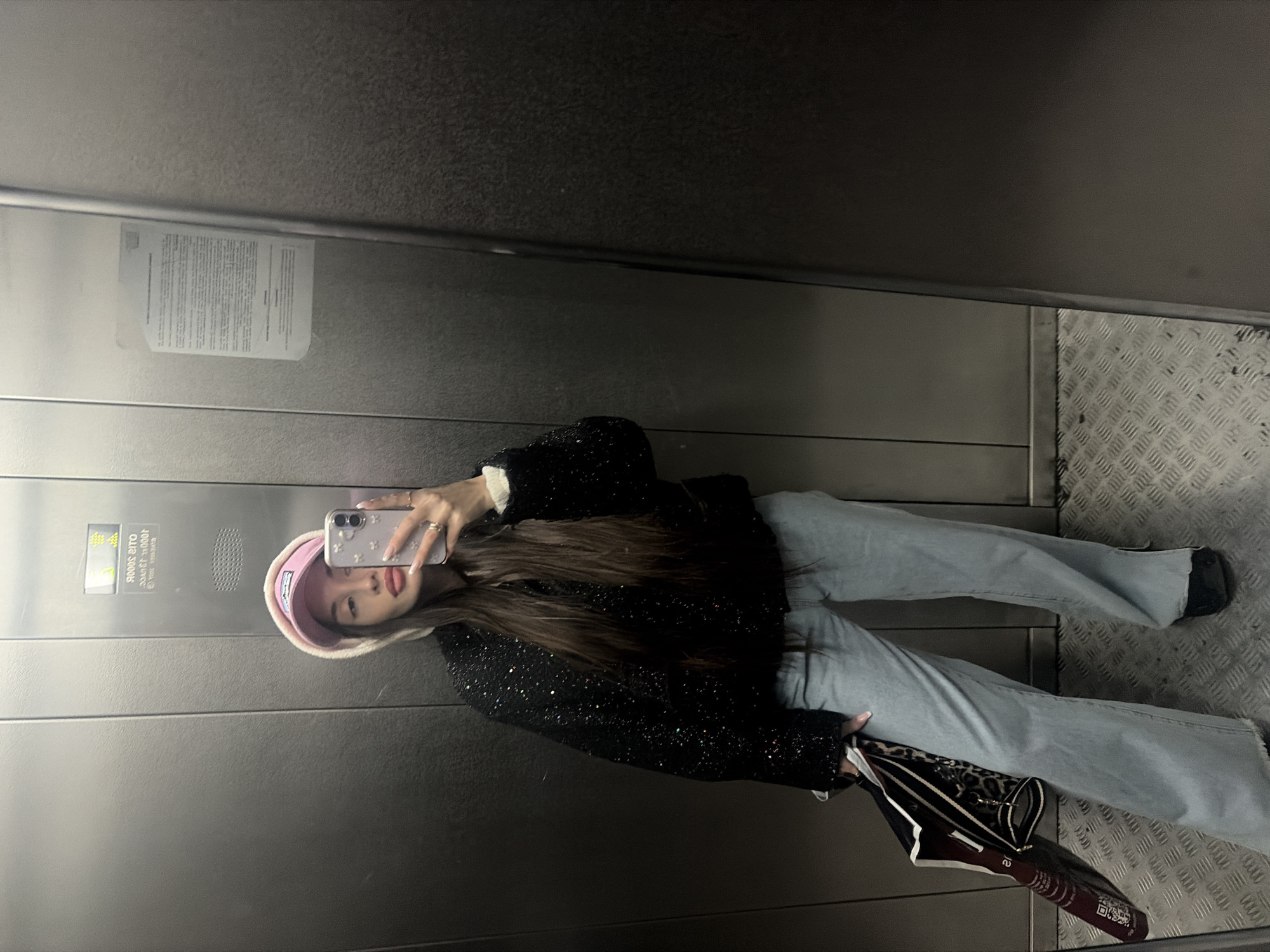 A woman is taking a selfie in an elevator. She is wearing a pink hat, a black jacket with sparkly details, and light blue jeans. She is holding a phone with a floral case. In the background, there is a metal wall with a warning sign and a metal floor with a textured pattern. An umbrella is lying on the floor next to her. The location is likely in Saint Petersburg, Russia, but the image does not contain enough clues to make that determination.