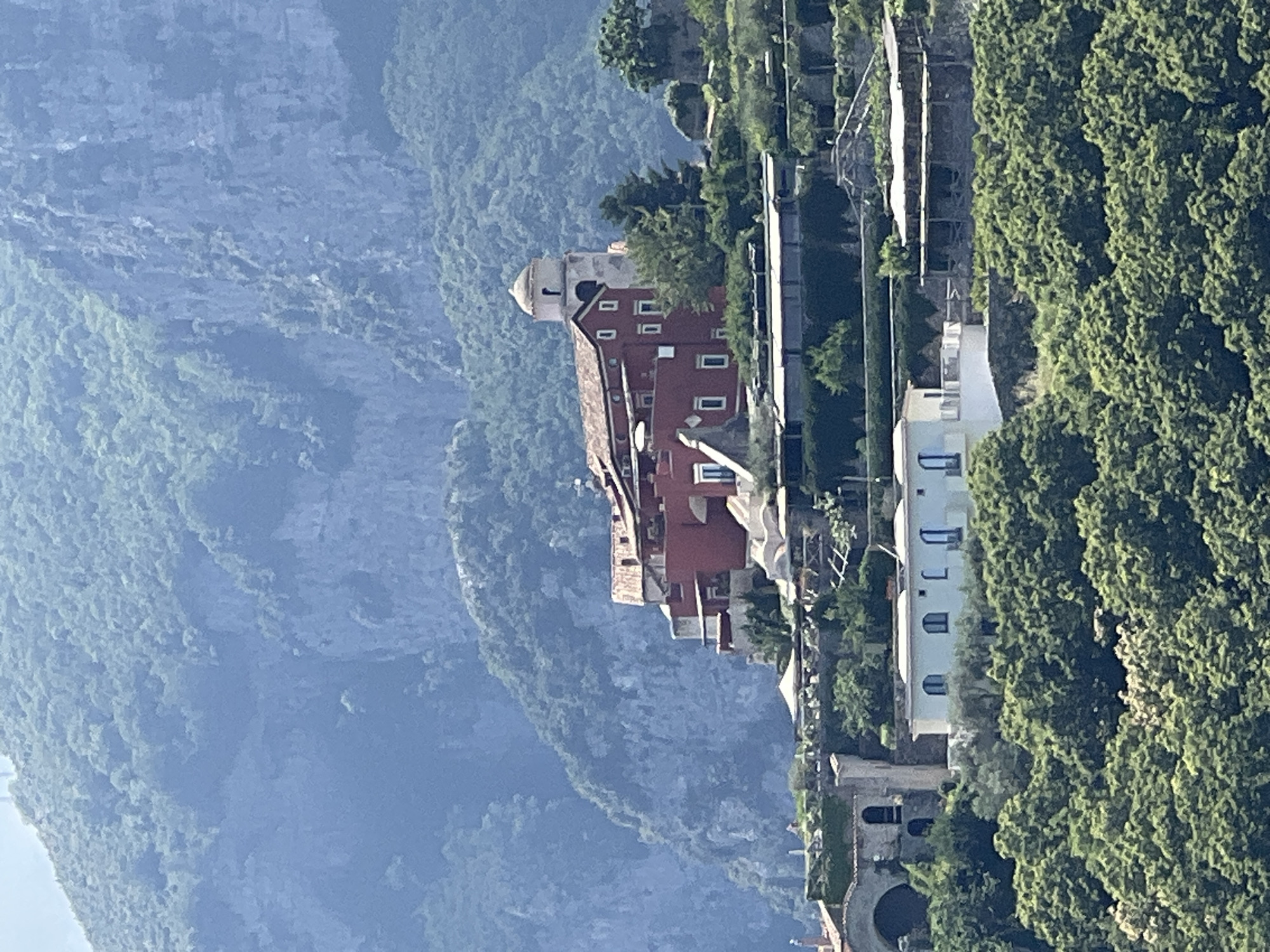 The image depicts a sunlit, terraced hillside in Ravello, Italy, characterized by lush green foliage and several buildings constructed with a mixture of terracotta, white, and stone materials. Dominating the mid-ground is a prominent red building with a white-domed bell tower, suggesting it might be a church or monastery. This structure is integrated into the hillside, with several levels of patios, balconies, and white awnings visible. Below this, a long, low white building with arched windows runs horizontally across the scene, surrounded by dense greenery. The background is comprised of a hazy, steep, mountainous landscape covered in trees. No people are visible in the image. The overall impression is one of a picturesque, historic Italian coastal town.