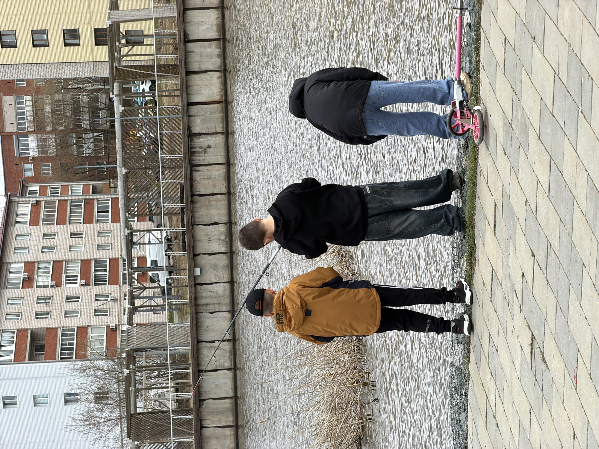 Three individuals are positioned along a water's edge. One person, wearing a mustard-colored jacket, black pants and a baseball cap, holds a fishing rod. Another individual, dressed in a black jacket and dark jeans, leans forward observing. The third person, wearing a black jacket and light blue jeans, stands beside a pink scooter. The backdrop includes urban structures, likely apartment buildings, and a retaining wall. The location is suggested to be the city of Votkinsk, Russia. The weather appears overcast.