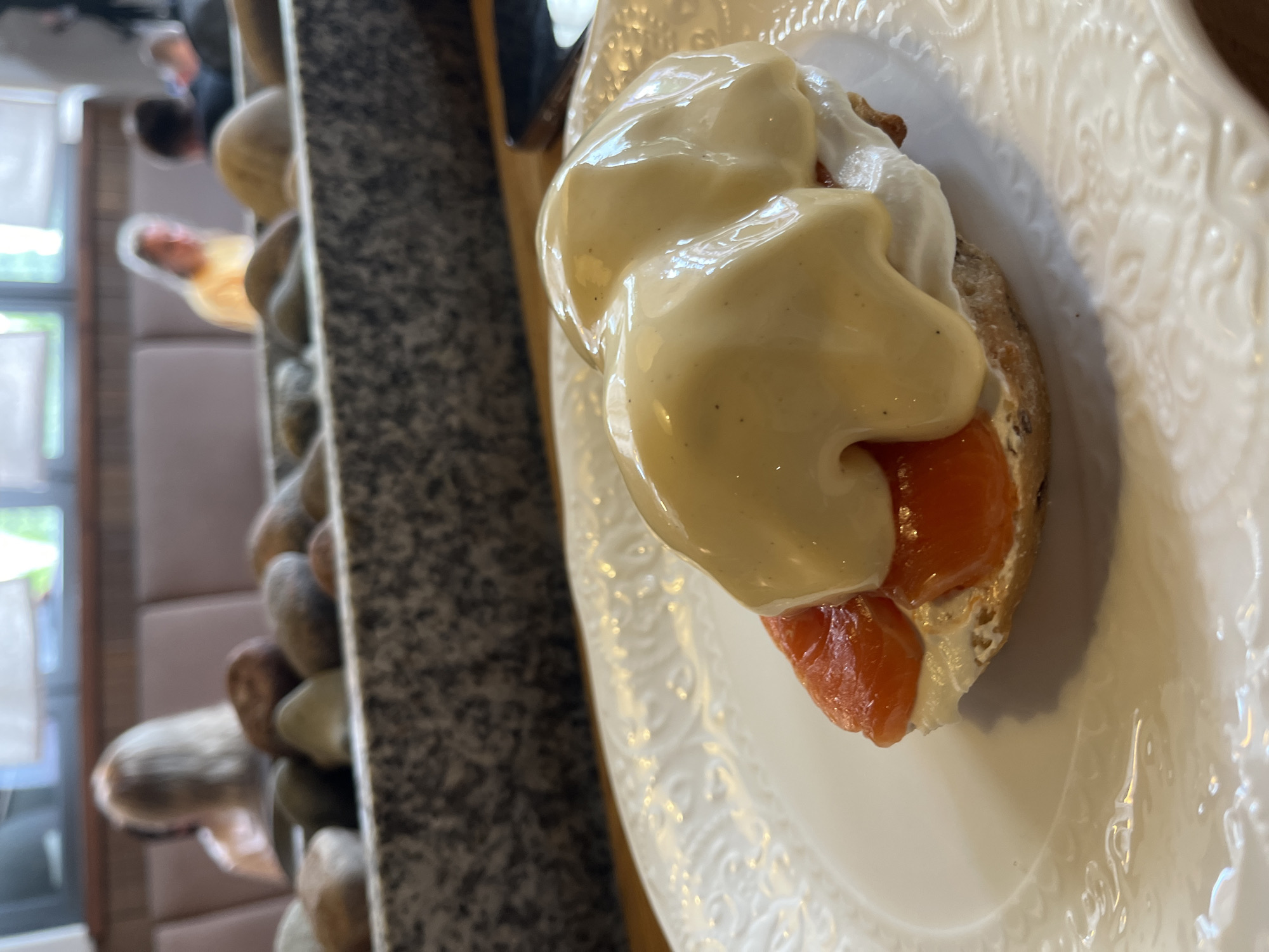 A close-up, angled shot shows a dish of salmon eggs Benedict on a white, textured plate. The dish features a toasted English muffin topped with cream cheese, slices of smoked salmon, and a generous serving of hollandaise sauce. The background is blurred, but shows people seated in a restaurant with a decorative rocky partition in the foreground. The setting appears to be a restaurant in Zelenogradsk, Russia.