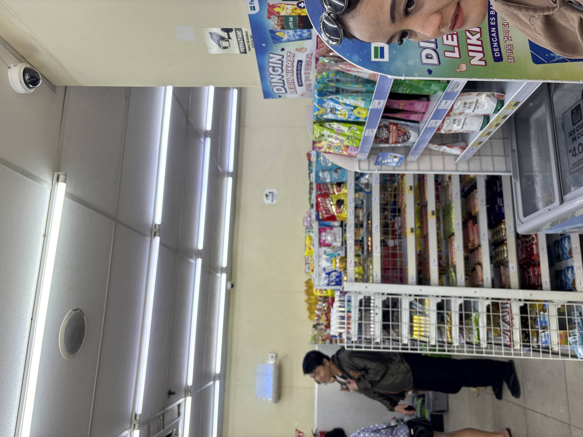 The image captures an indoor scene, likely a convenience store in North Jakarta, Indonesia. Fluorescent lights illuminate the ceiling, which also features a small, circular speaker. A security camera is visible in the upper right corner, mounted on the ceiling. In the foreground, a woman wearing sunglasses and a brown top is partially visible on the right side, looking towards the right. In the middle ground, a man in a green jacket and black pants is standing, facing left, with his hand on his chest. He appears to be looking at shelves stocked with various packaged goods. To the left of the man, a person wearing a blue and white polka dot shirt and a black purse is mostly out of frame. Shelves filled with merchandise, including bags of snacks and drinks, are prominent throughout the scene. A large advertisement featuring a cooler with drinks and text in Indonesian and Korean is displayed on the right. The floor is tiled.