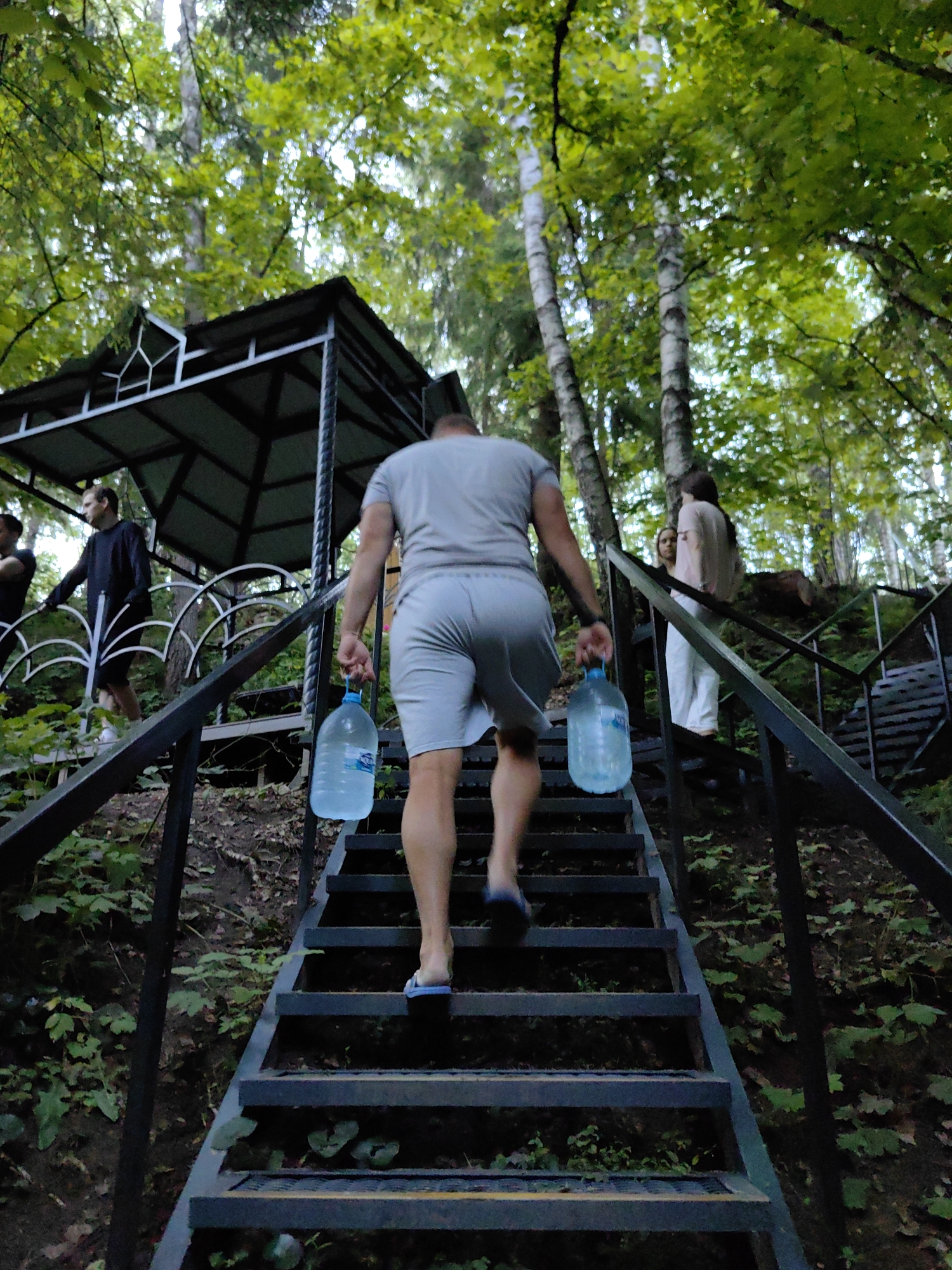 The image depicts an outdoor scene, possibly in the vicinity of Дзямідавіцкі сельскі Савет, Belarus, given the dense foliage and the presence of a wooden structure that resembles a gazebo or shelter, common in such rural or park-like settings. A man, viewed from behind, is ascending a metal staircase while carrying two large, clear plastic water bottles, one in each hand. He is wearing a light-colored t-shirt and grey shorts, and flip-flops. Other individuals are visible further up the stairs, one man in a dark long-sleeved shirt and shorts, and another person in a light-colored top and white pants. The staircase is bordered by dark metal railings and is surrounded by lush green vegetation, with trees forming a canopy overhead, suggesting a wooded or natural environment. The lighting suggests it is daytime, with dappled sunlight filtering through the leaves.