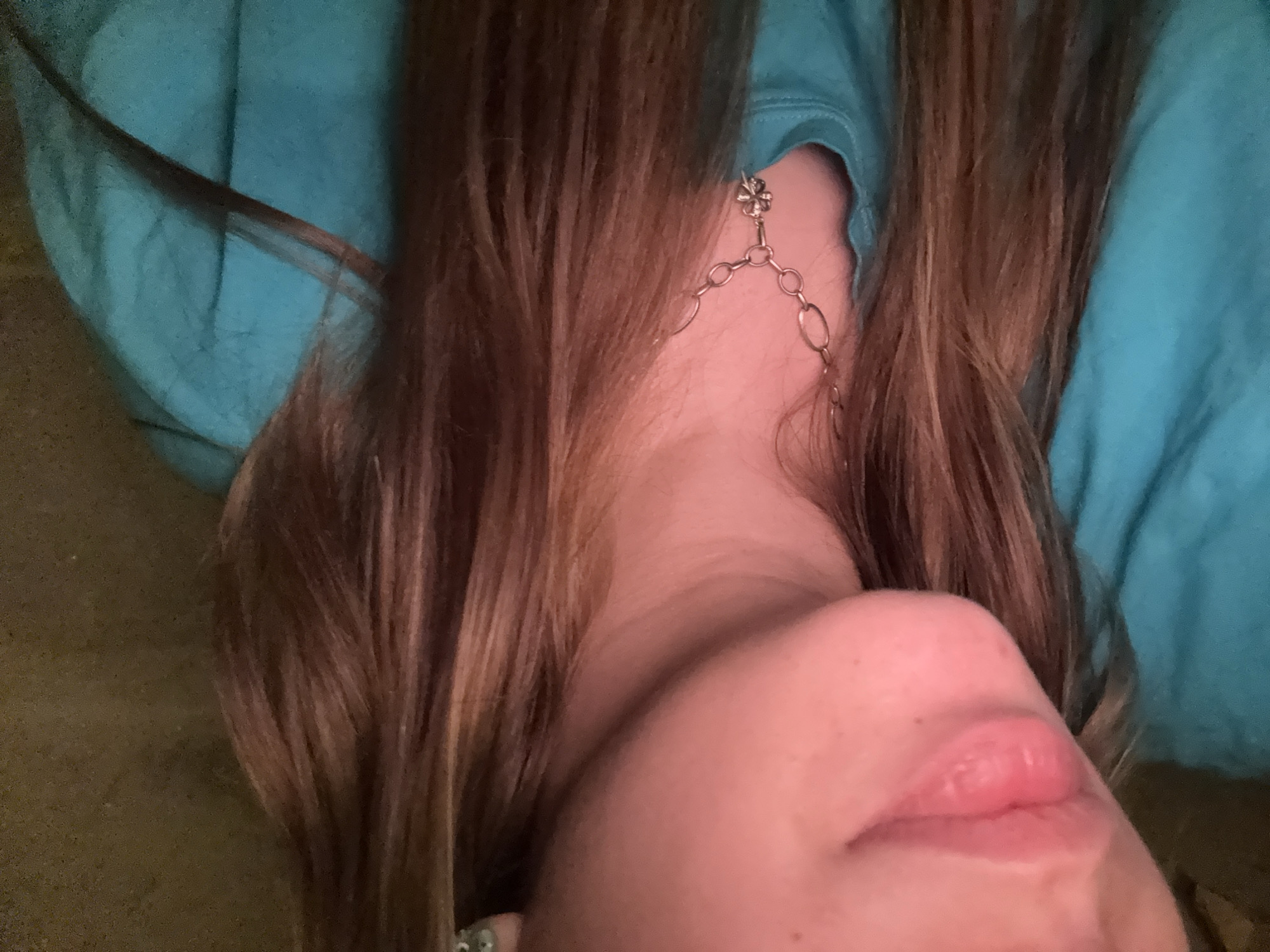 A close-up, angled portrait captures the lower portion of a person's face, neck, and hair. The subject's head is tilted back, revealing their chin, moisturized lips, and neck. Long, brown hair, streaked with auburn highlights, frames the face and spills around the neck area. The person is wearing a teal or turquoise garment, likely a sweatshirt, visible on the left side of the frame and around the collar.
A notable detail is a unique piece of jewelry adorning the person's left ear (viewer's right). It appears to be an elaborate earring or ear cuff, featuring a small, silver-toned decorative element at the top, possibly resembling a cross or a four-leaf clover. From this top piece, delicate, linked chains extend downwards, tracing the curve of the neck. The lighting is soft and diffused, highlighting the skin's texture and the sheen on the lips. The background is a muted, earthy brown, possibly a piece of furniture or carpet, suggesting an indoor setting. The image is tightly cropped, focusing on these intimate details rather than a full view of the subject or surroundings. There is no specific activity depicted, nor any visible text.
