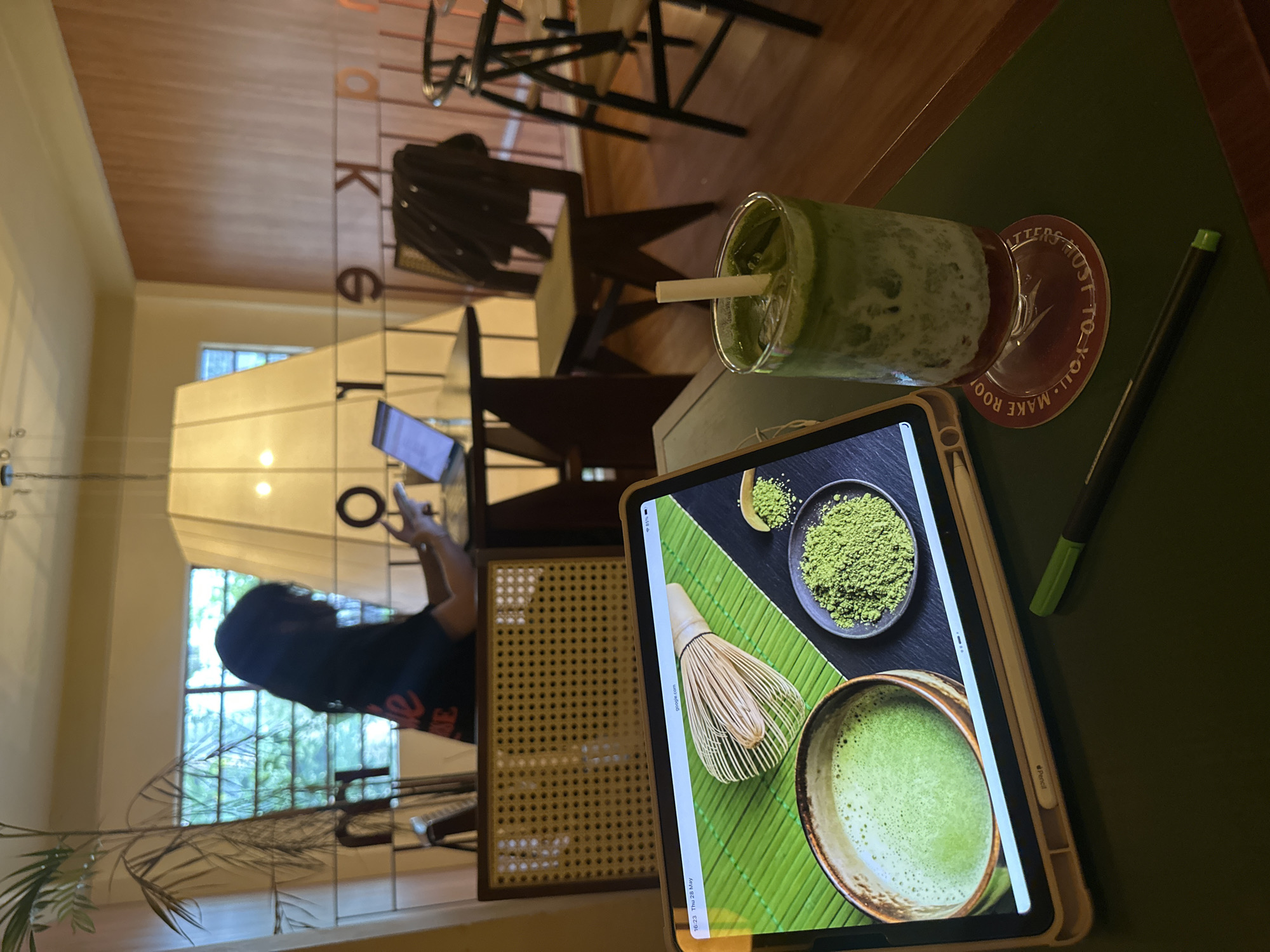 The image shows a scene at a cafe in Thủ Đức, Vietnam. A person is partially visible in the background, sitting near a window and using a tablet. In the foreground, a tablet displays a detailed image of matcha-related items, including matcha powder, a whisk, and a bowl of matcha. Beside the tablet, there's a glass of matcha drink with a straw, resting on a coaster with text. A stylus pen lies nearby. The background shows a cafe interior with wooden furniture and a view of greenery outside the window.