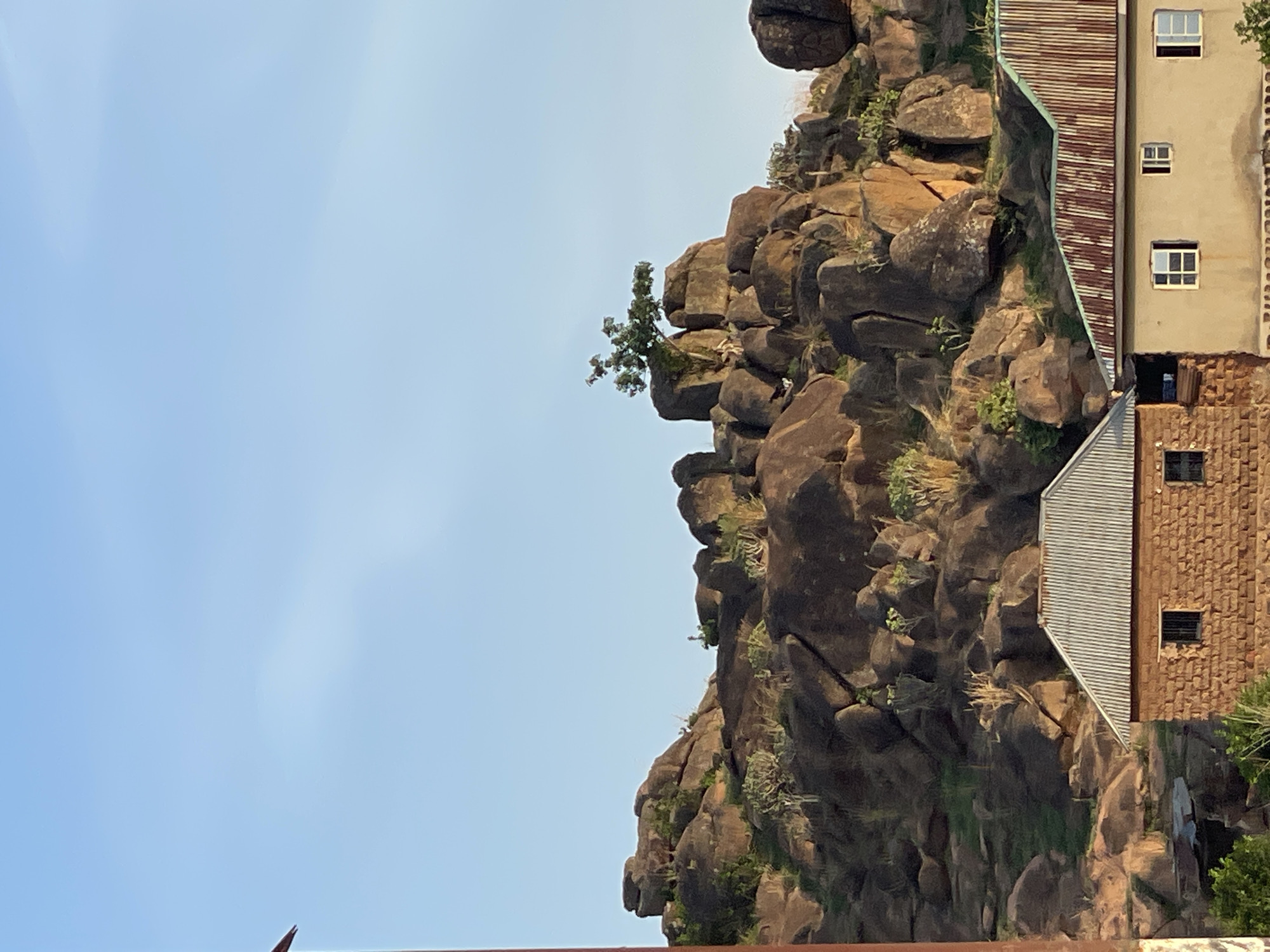 The image shows a rocky outcrop adjacent to a building in Barikin Gidar Kanat, Nigeria. The outcrop is composed of large, dark brown boulders and sparse vegetation. A small tree is growing on the upper section of the rock formation. The building is directly next to the rock, with what appears to be a roof section visible with a corrugated metal finish, and a multi-story side with windows. The sky is light blue and appears clear.