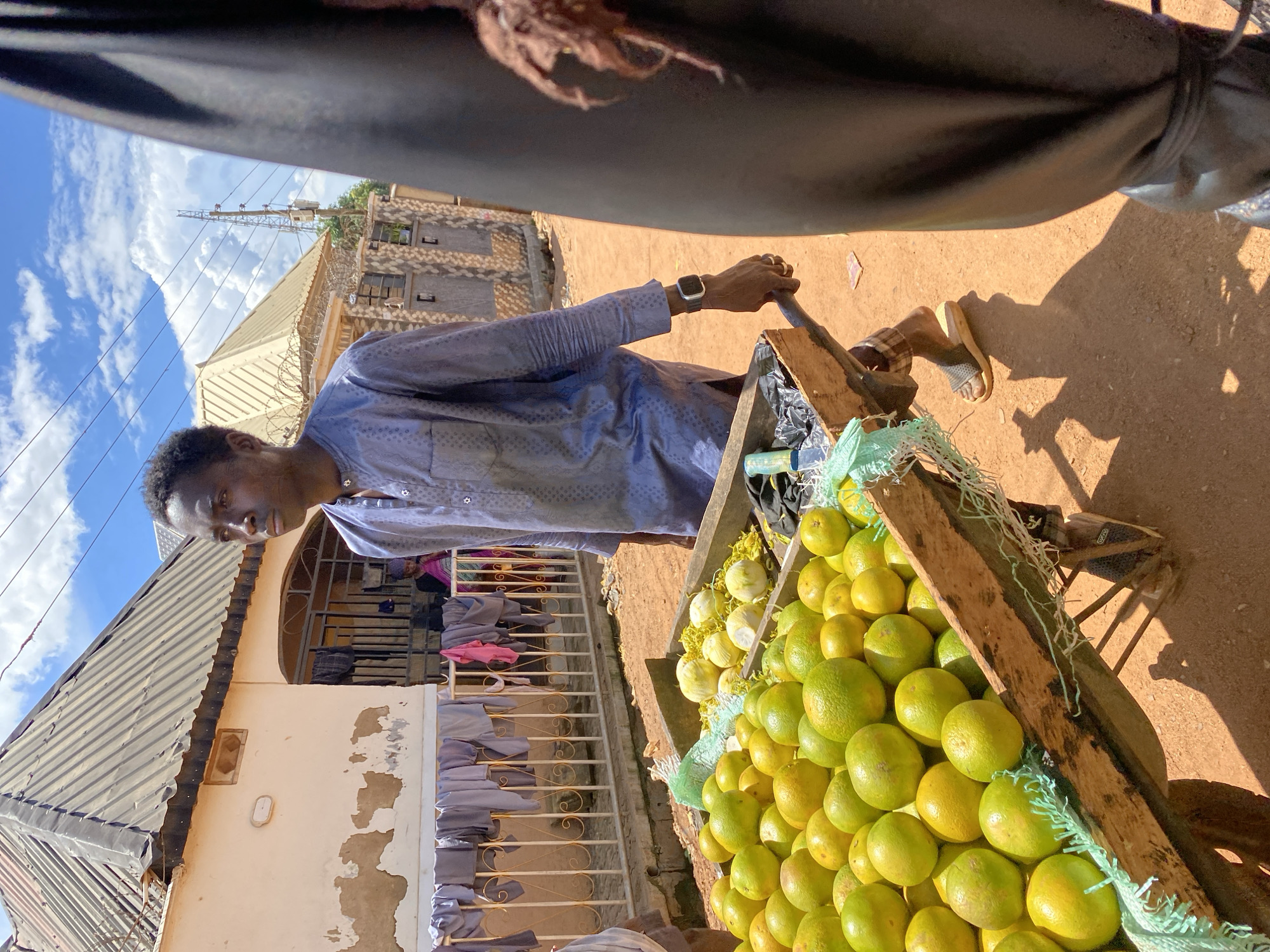 An outdoor, eye-level, medium shot captures a young Nigerian man from Bukuru, Nigeria, pushing a wooden cart filled with green oranges. The man, dressed in a blue traditional outfit and wearing a watch, is seen from the chest down, his face turned to the left as he pushes the cart. The cart is laden with a generous amount of ripe, green oranges, some of which are partially covered by a light green mesh. Behind the man and the cart, a residential building with a corrugated metal roof is visible, with laundry hanging on a railing in front. The ground is a reddish-brown dirt, typical of the region. The sky above is blue with scattered white clouds, indicating a sunny day. The scene appears to be a street or market setting in Bukuru, Nigeria.