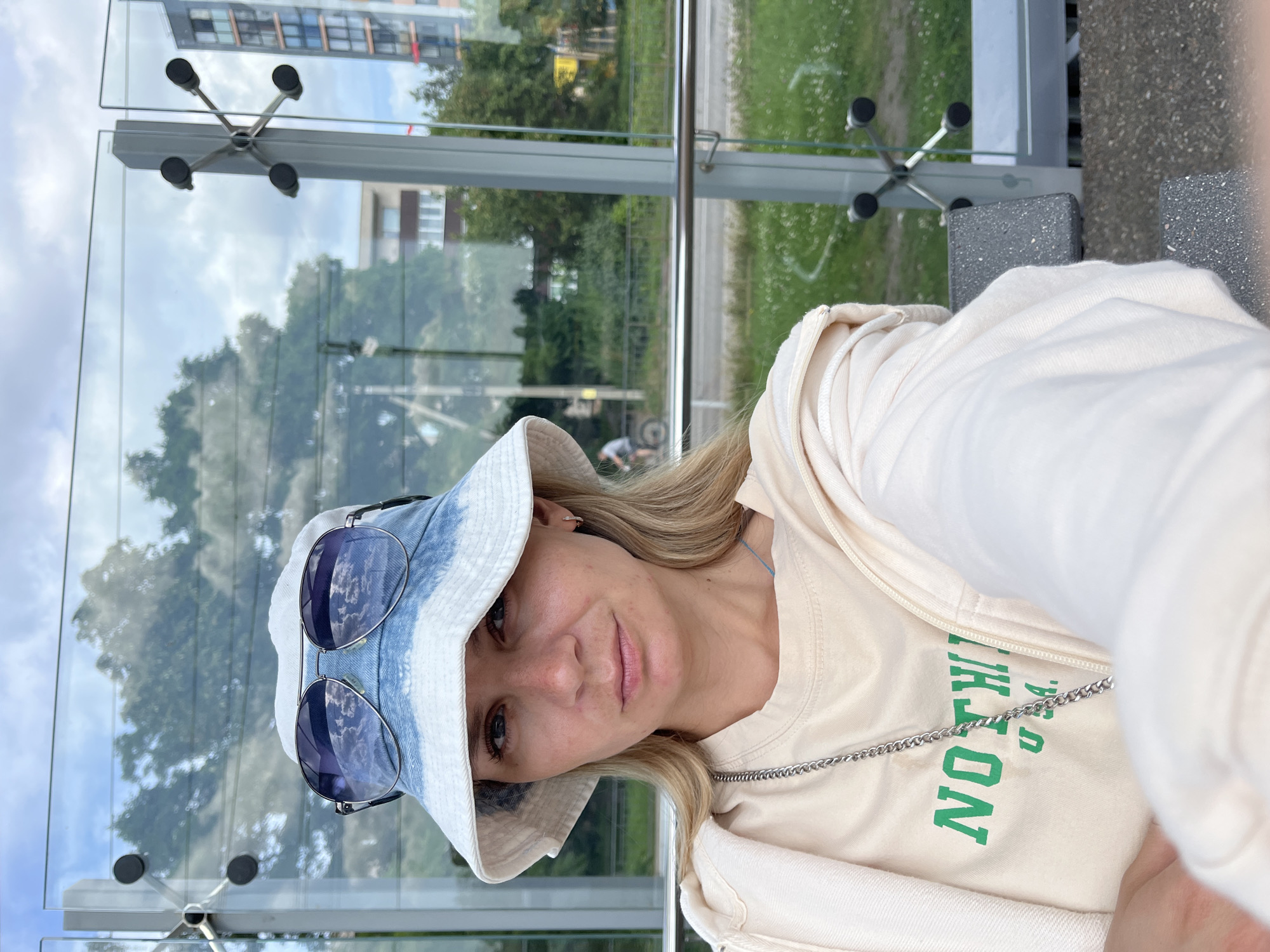 A young woman with blonde hair, wearing a denim-accented bucket hat and sunglasses, is taking a selfie. She is dressed in a light-colored t-shirt with green lettering that reads "NOTHING U.S.A." and a cream-colored zip-up hoodie. She is seated on a gray bench, which appears to be part of an outdoor waiting area, possibly a bus stop or a public transport shelter. The shelter has large glass panels that reflect the cloudy sky and trees, with a glimpse of buildings and a road in the background, suggesting an urban or suburban setting. A person on a bicycle is visible in the distance. The location is identified as Pionersky, Russia.