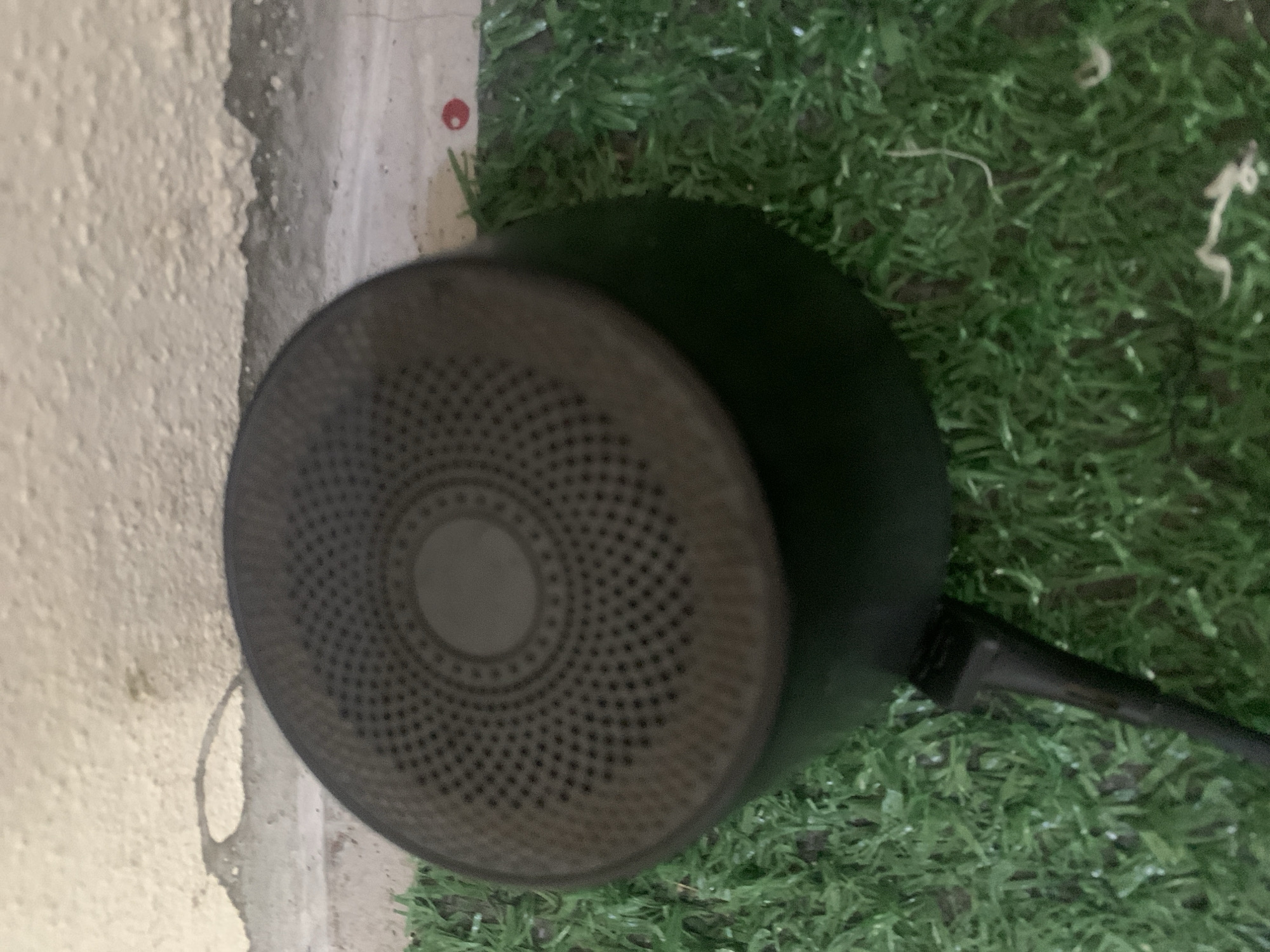 A close-up, eye-level shot shows a dusty, dark-colored portable speaker resting on a patch of artificial grass. The speaker's top surface is textured with concentric circles of small perforations and a central, slightly raised circular area. A black cable extends from the bottom of the speaker and disappears into the grass. Behind the speaker, a textured, light-colored wall with some discoloration is visible. The scene is set against the backdrop of Jauro Yinu, Nigeria.