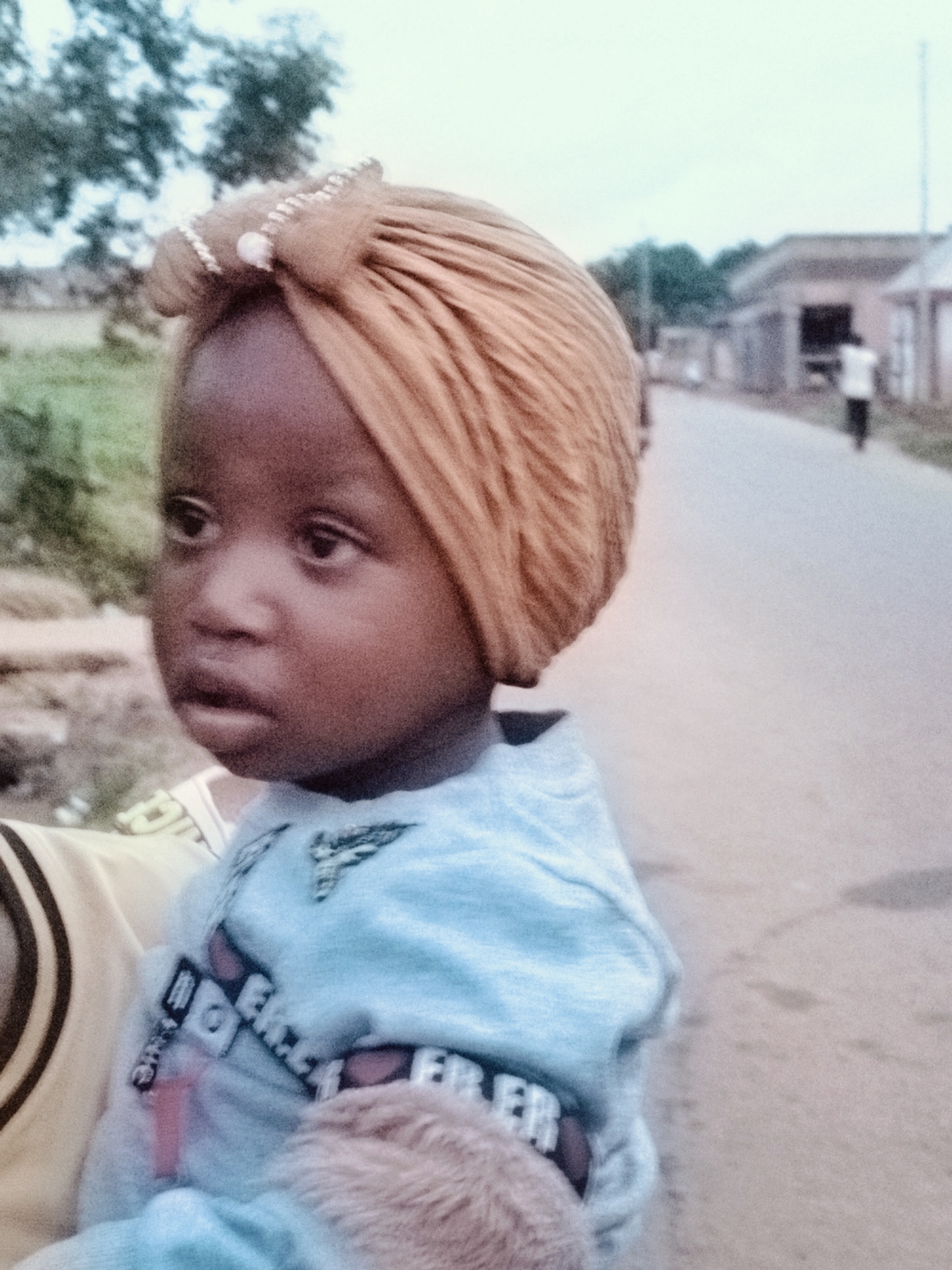 A young child, likely a toddler, is the primary subject of this image, captured in a close-up portrait. The child is facing the right side of the frame, with their head turned slightly towards the viewer. They are wearing a light blue long-sleeved shirt adorned with a white embroidered emblem on the chest, and a decorative band with text and possibly images on the sleeve cuff. A tan-colored turban, embellished with small white beads and a central light pink bead, is worn on the child's head. The child's facial features are dark, with expressive eyes looking slightly off-camera. The background is blurred, indicating a shallow depth of field. To the right, a paved road is visible, with a person walking in the distance. Buildings with roofs are also present in the background, suggesting an outdoor urban or semi-urban environment. The overall lighting suggests an overcast day. The context provided specifies the location as the city of Du, Nigeria, implying this scene takes place within that locale.