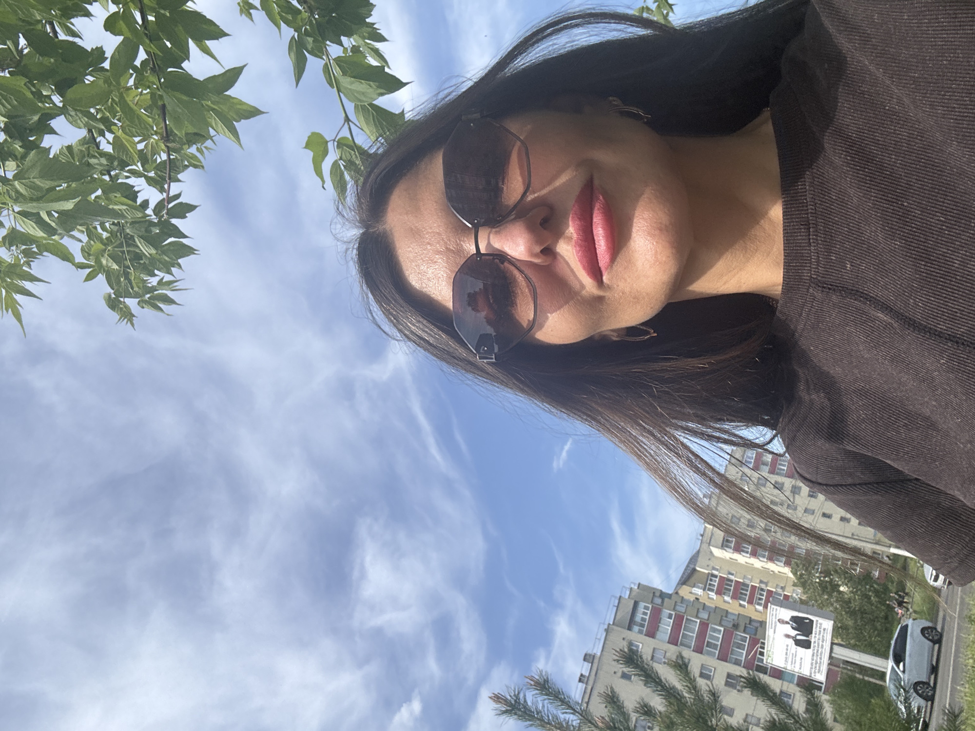 The image is a selfie of a woman with long, dark hair wearing sunglasses and a dark shirt. She appears to be outdoors. The background shows a partly cloudy blue sky, greenery including trees and bushes, and a multi-story building with red and white accents. A billboard is visible near the building. The building suggests a location in an urban area.
Based on these details, the location is likely in Chaikovsky, Russia as indicated in the prompt. The architecture of the building, presence of trees, sky, and billboard are all consistent with what one might expect in such a location.