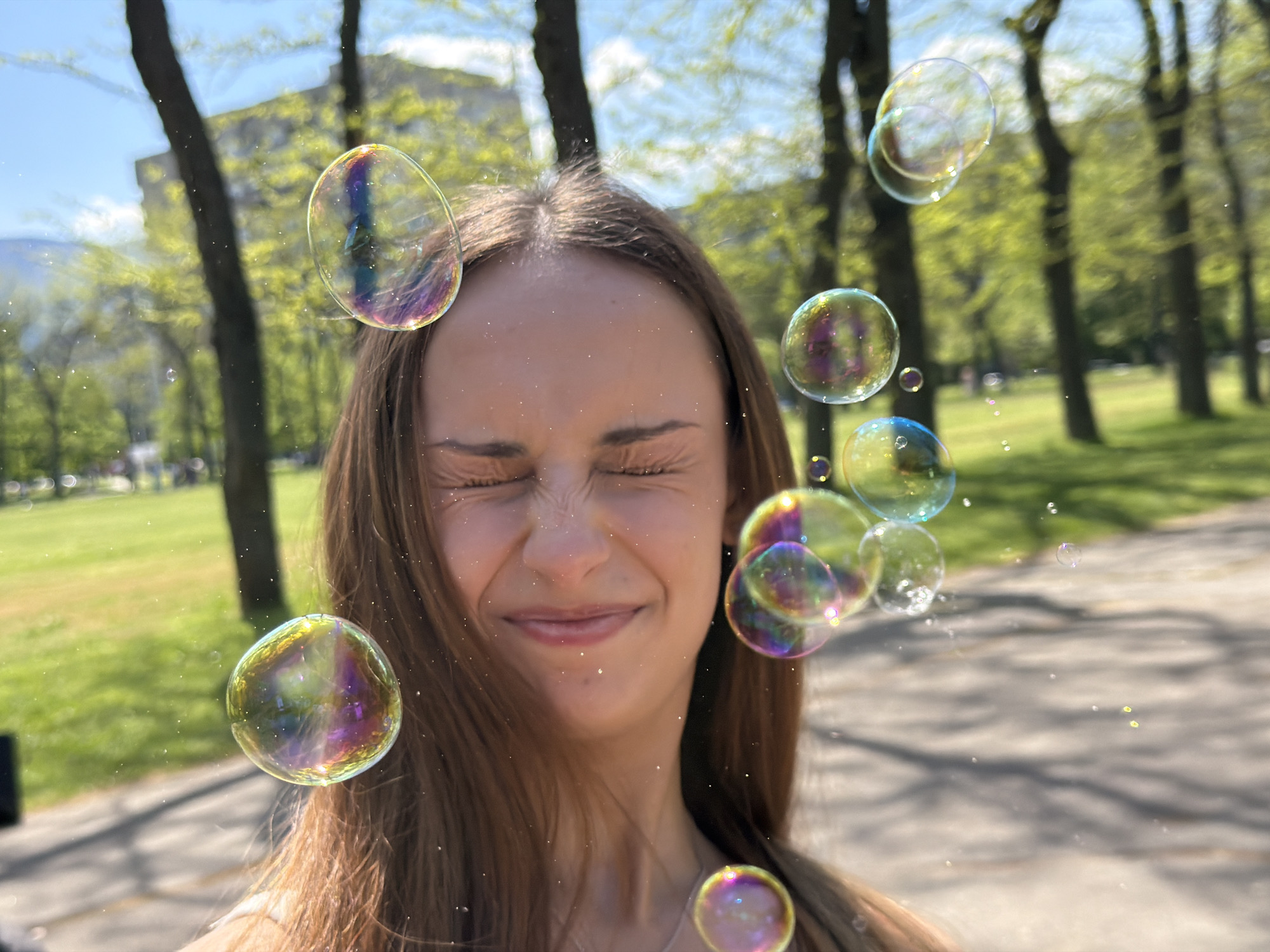The image depicts a young woman, likely in her late teens or early twenties, with long brown hair, standing outdoors on a sunny day. Her eyes are closed, and she has a slight smile on her face. Soap bubbles are scattered around her, some reflecting light and showing distorted images of the surroundings.
The background shows a park setting with trees and a building in the distance, suggesting an urban environment. The location is most likely in the city of Sofia, Bulgaria, based on metadata analysis, as the image shares similarities with images found there. A sidewalk is visible under the trees. The overall scene is playful and carefree.