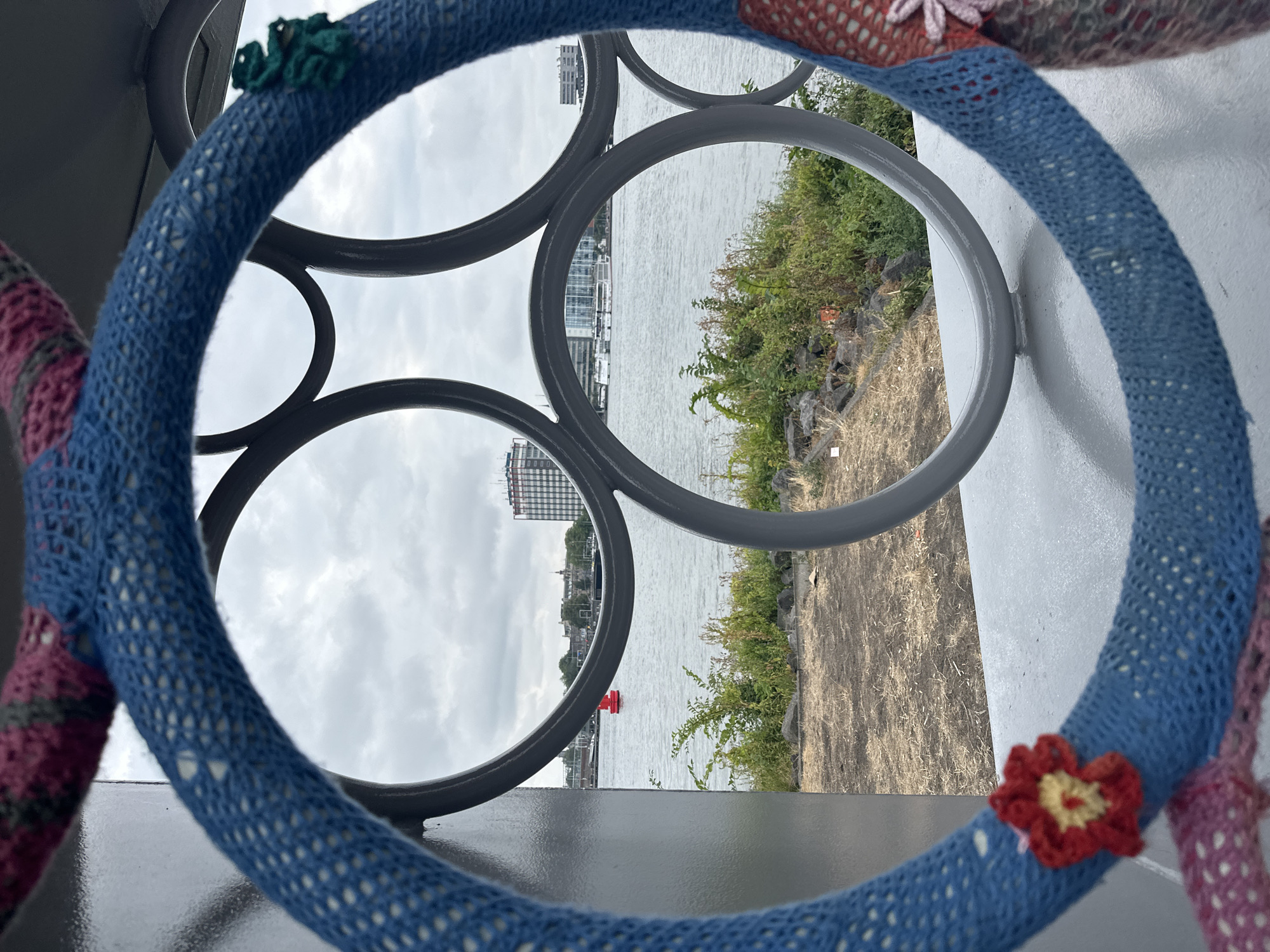 A medium shot captures a view through a knitted circular sculpture onto a waterfront scene in Amsterdam, Netherlands. The sculpture itself is decorated with crocheted flowers, one red and yellow on the left, and a green embellishment on the upper right. The background shows a body of water, with a red buoy visible in the distance. Along the waterfront, there is a grassy bank with some rocks, and beyond that, buildings and a cloudy sky. The overall impression is one of an urban landscape softened by artistic embellishments.