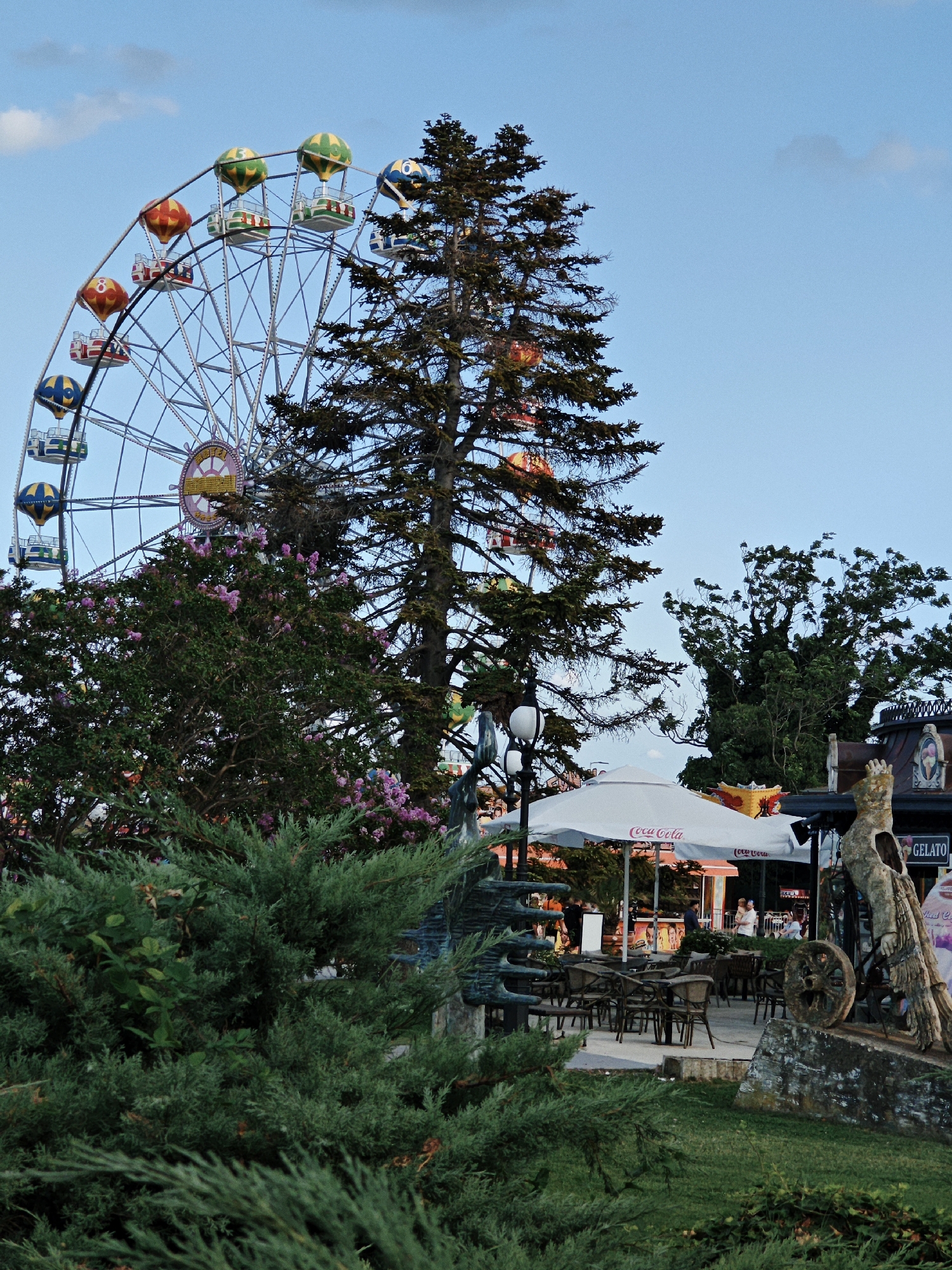 An outdoor Ferris wheel amusement park scene is depicted under a bright blue sky with scattered clouds. The Ferris wheel, with colorful gondolas resembling hot air balloons, dominates the left side of the frame, partially obscured by a tall evergreen tree. To the right of the Ferris wheel, a restaurant or cafe area is visible, featuring white umbrellas with "Coca-Cola" logos, tables, and chairs. Several people are present in the background, appearing to be patrons or staff. A statue or sculpture of a person with a wheel is situated in the foreground on the right, next to a patch of grass. Lush green bushes and flowering plants are in the foreground, adding a natural element to the amusement park setting. The lighting suggests it is late afternoon or early evening.