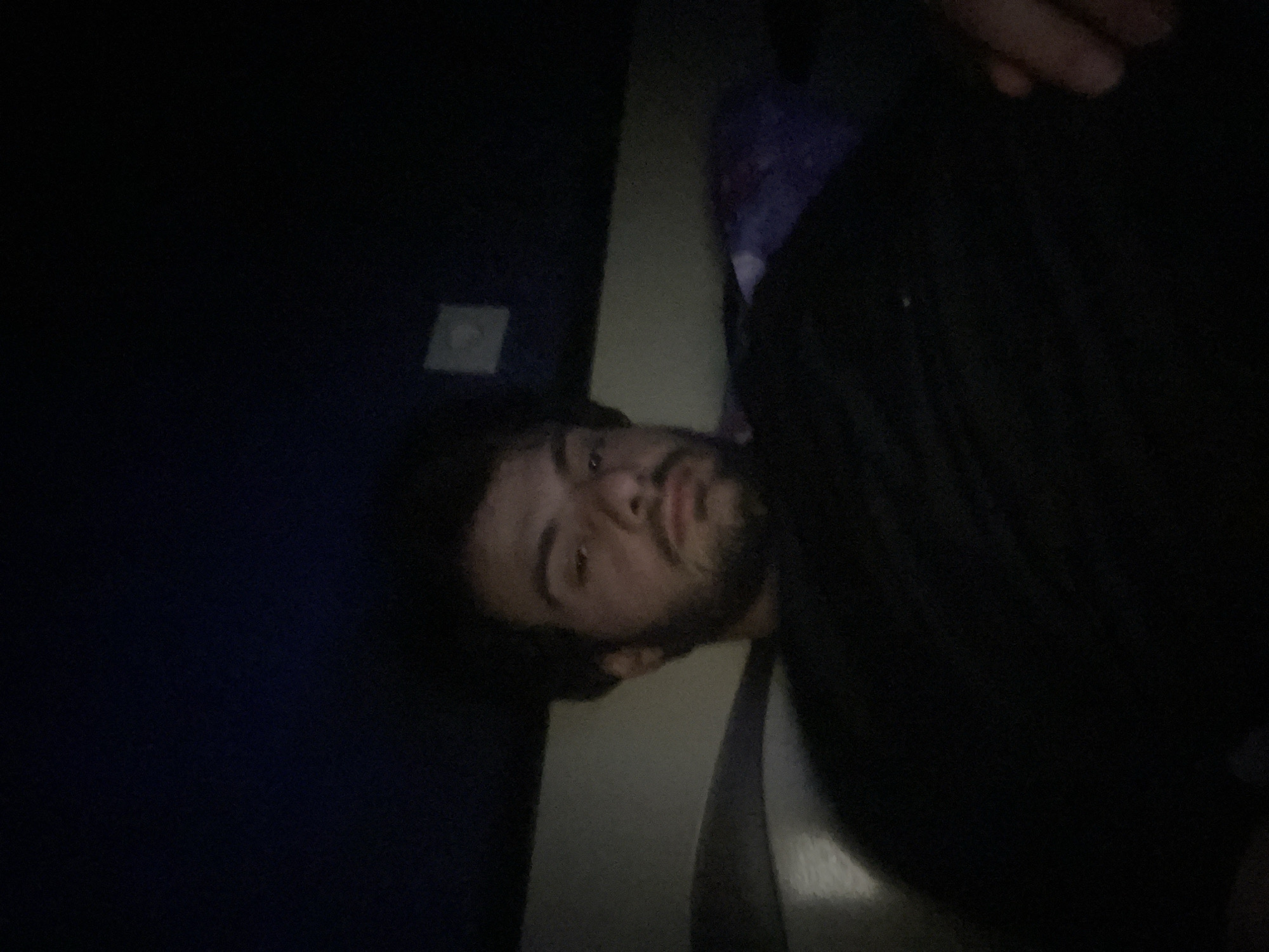 A dimly lit, low-angle, medium shot captures a young man with dark, curly hair and a beard from the chest up. He is looking directly at the camera with his eyes partially closed. He is wearing a black t-shirt. The background consists of a dark, textured surface that appears to be a wall or ceiling, with a small, square light fixture visible on the upper right. Below this, a lighter-colored surface suggests a wall or partition, and to the right, a section of dark fabric with a purple pattern is partially visible. The overall impression is of an indoor setting, possibly a room or a cinema, given the low light and the presence of what might be seating. There is no indication of Tbilisi, Georgia, in the image itself.