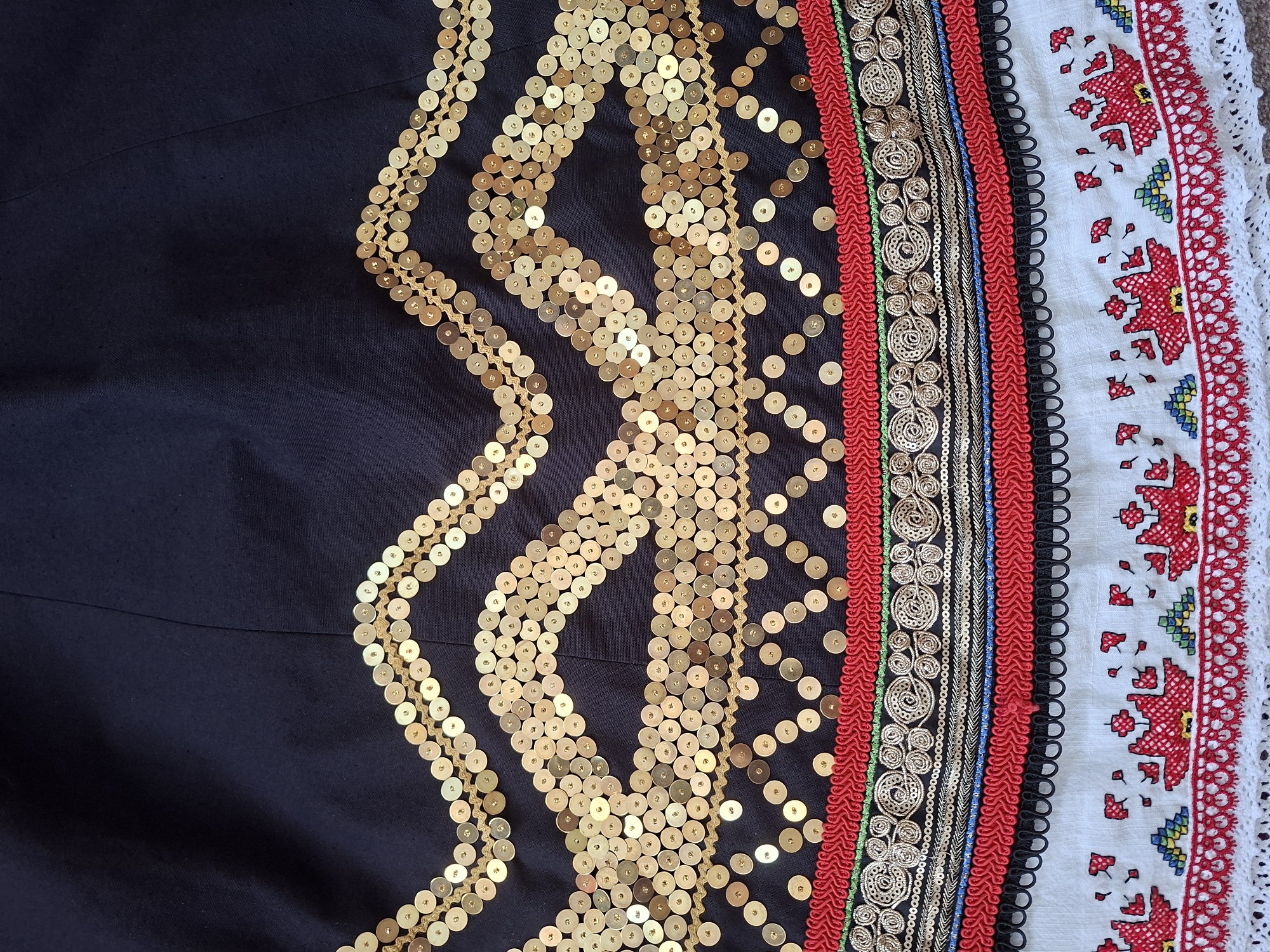 The image shows a close-up view of a dark fabric, possibly a skirt or dress, adorned with intricate embellishments. The top portion features multiple wavy rows of gold sequins arranged on a tan string, creating a shimmering effect against the dark background. Below the sequins, there are several decorative bands of trim. One band is red with a woven textured pattern, followed by a thin green line. Next is a wider band with gold swirling patterns and small beads, then a thin band of black and gold cording, and another red band with a different woven pattern. A black trim with looped elements separates the embellished section from the bottom portion. The bottom part of the fabric is white and features embroidered red floral or folk art motifs, interspersed with small green, blue, and yellow geometric shapes. A white lace trim finishes the bottom edge. There are no people or environmental details visible, as the focus is entirely on the textile details. The location is Unknown, Unknown.