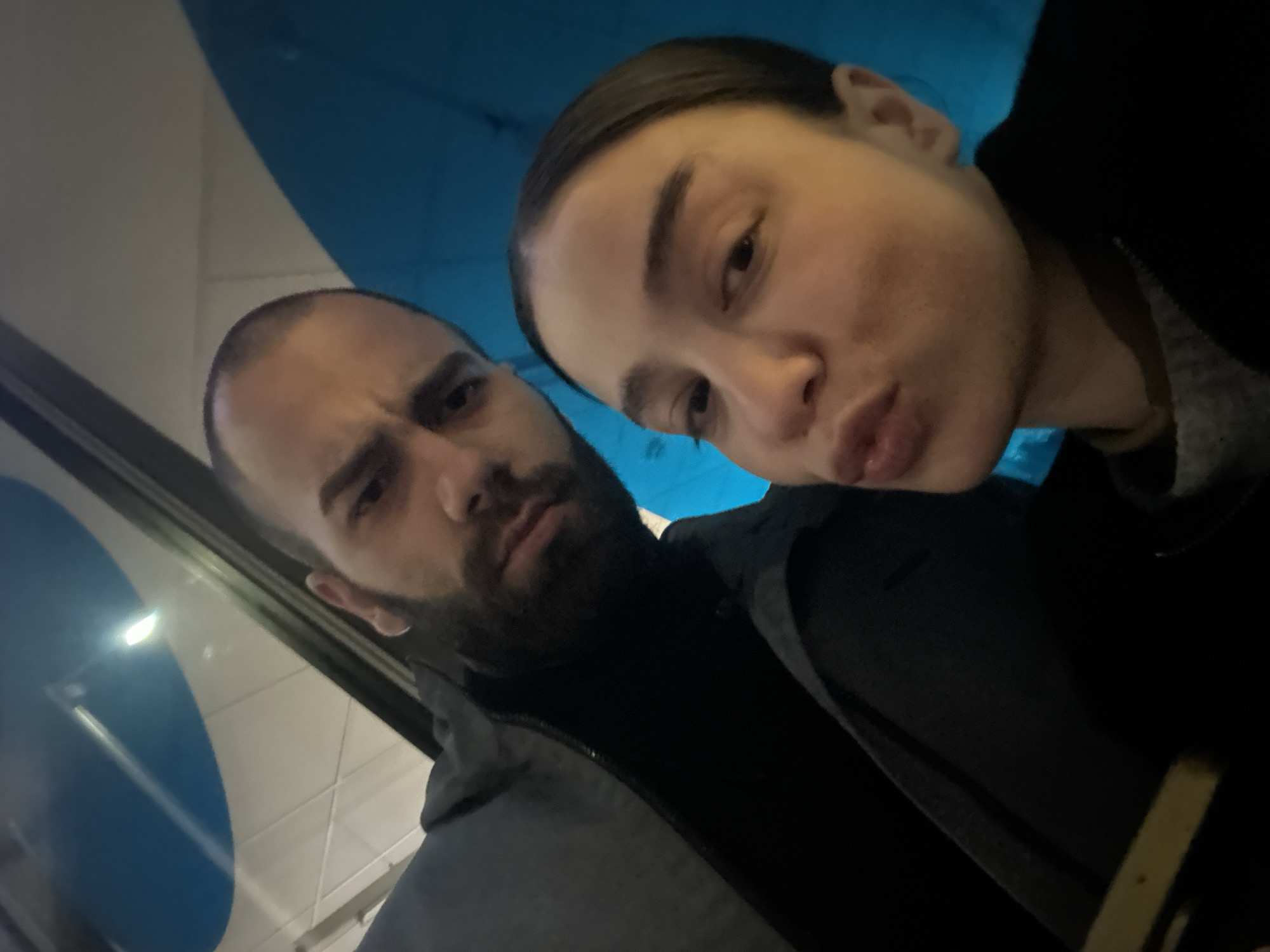 Two people are close to the camera, posing for a selfie. A man with a dark beard and a bald head is on the left, looking at the camera with a neutral expression. A woman with her hair pulled back is on the right, pursing her lips into a kiss. They are both wearing dark-colored clothing. In the background, part of a blue and white circular object is visible. The image is tilted, suggesting it was taken at an angle. Given the limited visual information, the location within the city of Sofia, Bulgaria, cannot be determined.