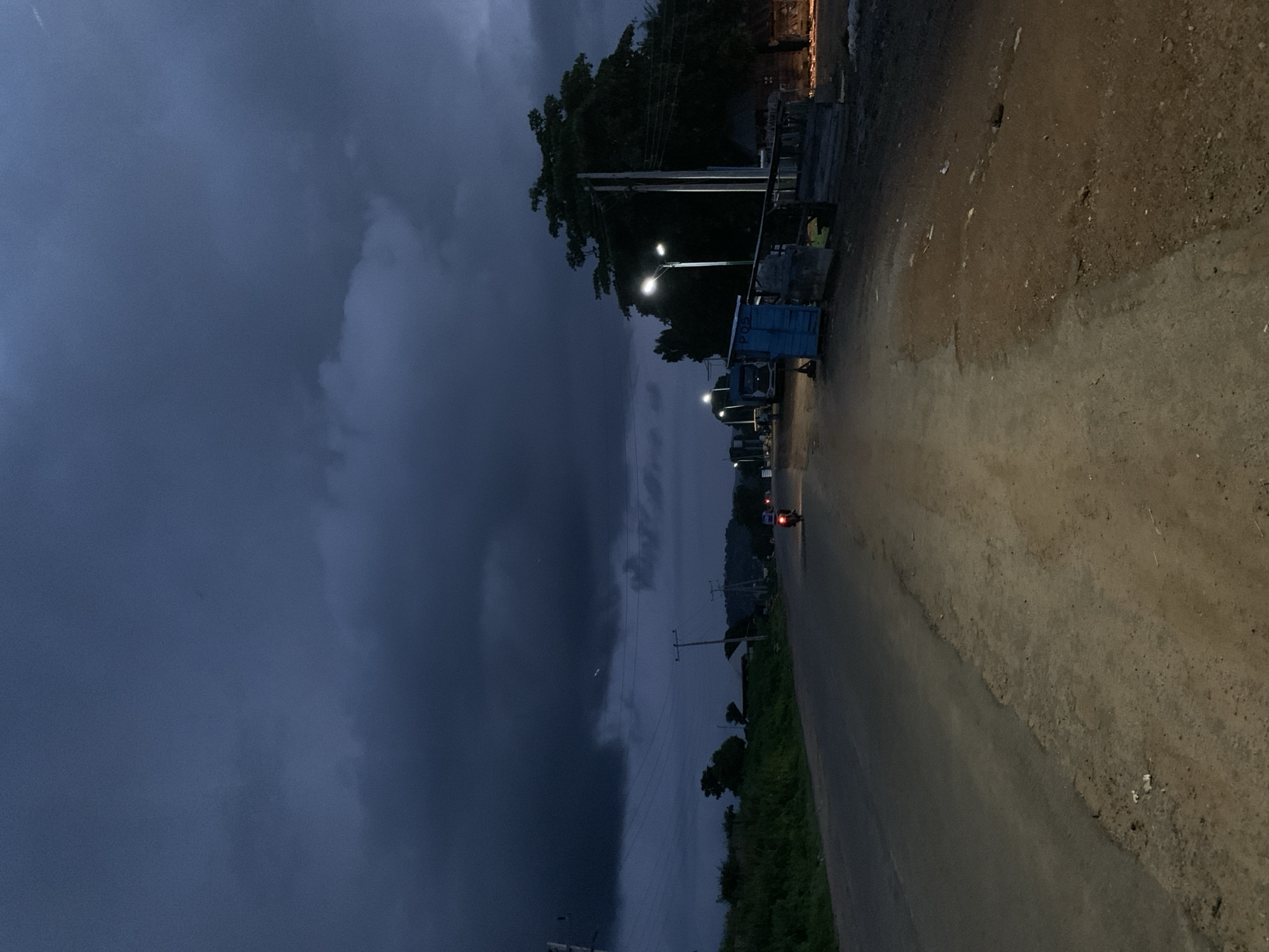 The image depicts a desolate, unpaved road in Kabba, Nigeria, under a heavily overcast, dark sky, suggesting impending rain or a storm. The road surface is a mix of asphalt and dirt, with loose gravel and debris visible. On the left side of the road, a grassy embankment slopes upwards, dotted with vegetation. On the right side, there are several structures, including a small blue kiosk with "PQZ" written on it, a wooden fence, and what appears to be a small building with a corrugated metal roof. Several streetlights illuminate the scene, casting a dim glow. A solitary motorcycle rider, illuminated by their headlight, is traveling away from the viewer down the road. In the distance, more streetlights and structures are visible, along with a hilly landscape. Power lines are strung across poles, adding to the rural or semi-urban atmosphere of the location. The overall mood is somber and quiet, emphasizing the isolation of the setting.