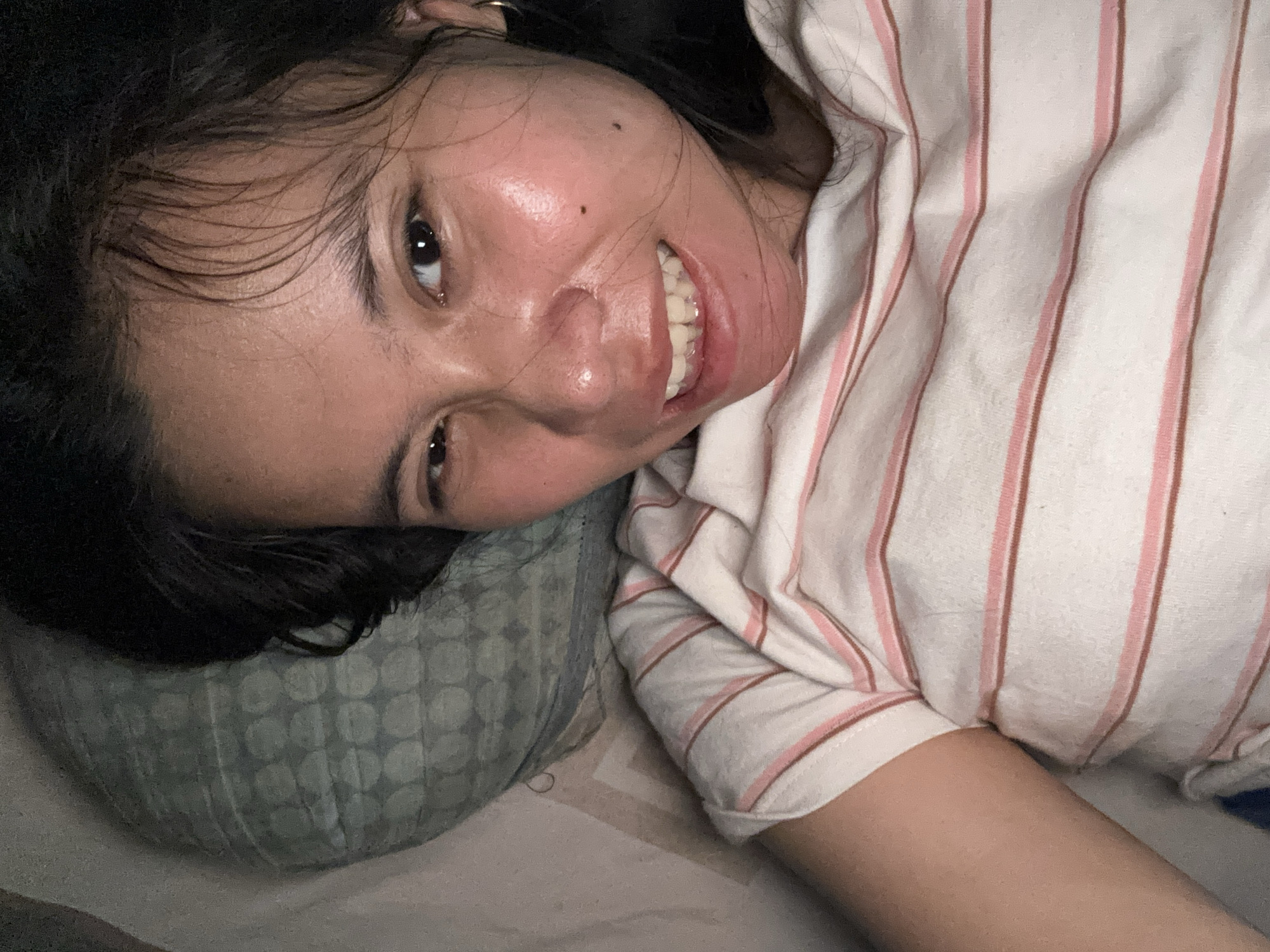 A young woman with dark hair and brown eyes is lying down on a pillow, her head tilted to the side. She is wearing a white t-shirt with thin pink and brown stripes. Her left eye is closed, and she is smiling with her teeth showing. The background consists of a pillow with a geometric pattern and a striped surface. The image appears to be taken indoors, likely in a bedroom. The location is East Jakarta, Indonesia.