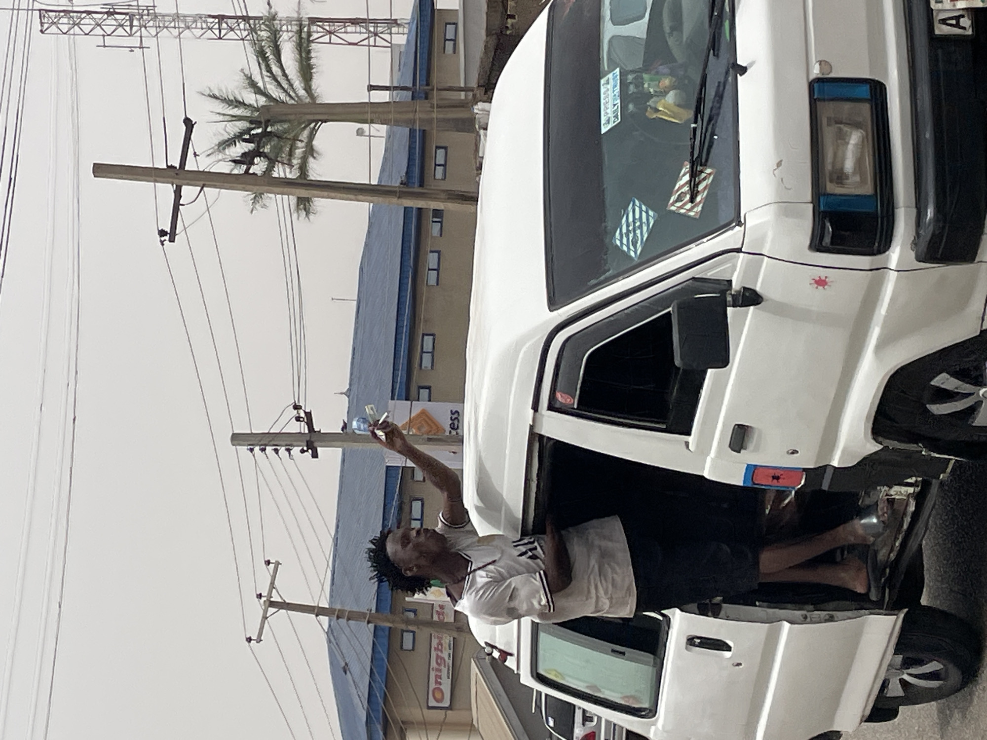A man stands in the open driver's side door of a white van, holding up what appears to be money. The van is parked on a street with power lines and poles overhead. Buildings are visible in the background, one with a sign that reads "Onigbinde". The location is in Jos, Nigeria.