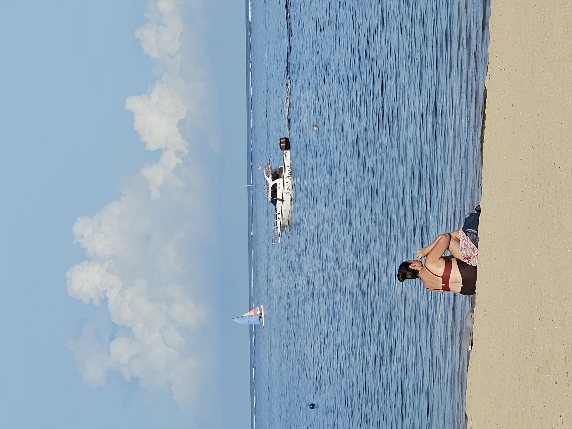 The image depicts a beach scene. On the right is a sandy beach where a woman is sitting, wearing a maroon-colored swimsuit top and dark shorts. She is facing the ocean. In the middle ground is the ocean, with a power boat labeled "Sweet Heart" and a sailboat in the distance. The sky on the left side is blue with large, puffy white clouds. The water is calm, with slight ripples visible. The location is Denpasar, Indonesia.