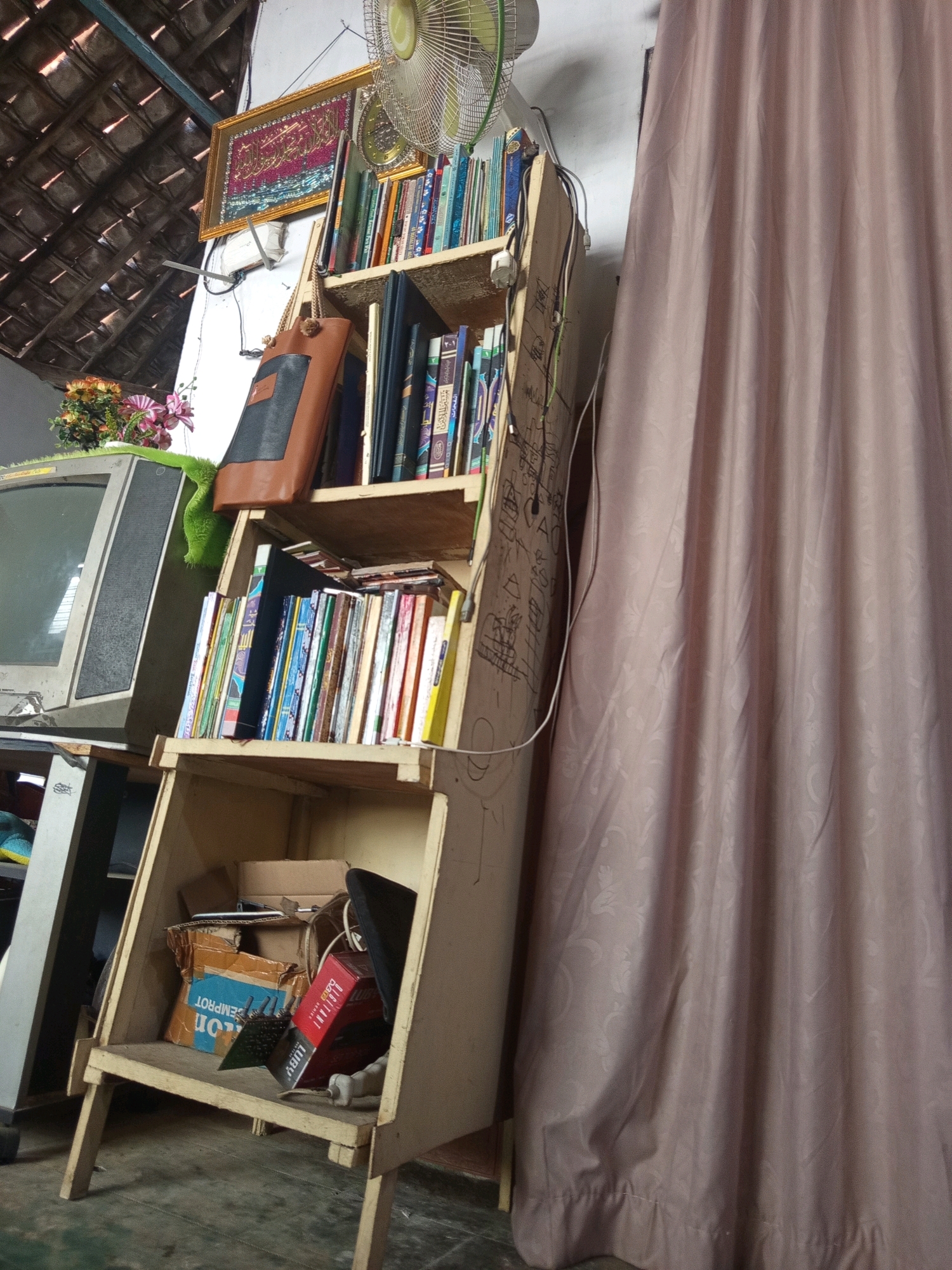 The image captures a dimly lit interior space, likely a room in a residential area of Ngablak, Indonesia, based on the visible architectural elements such as the tiled roof structure. Dominating the scene is a rustic, light-colored wooden bookshelf with multiple tiers. The bookshelf is laden with various items, predominantly books of different sizes and colors, neatly arranged on the shelves. On the top shelf, a framed artwork with Arabic calligraphy hangs on the wall behind the books, illuminated by natural light. Adjacent to this is a vintage fan mounted on the wall.
A brown leather bag with a black patch hangs from one of the shelves. On the lower shelves, more books are visible, interspersed with some loose papers and what appears to be an old television set on the left side of the frame. The bottom shelf of the bookshelf holds a collection of miscellaneous items, including cardboard boxes, a red object resembling a box of cigarettes, and other unidentifiable objects. A large, pale pink curtain with a subtle floral pattern drapes the right side of the image, obscuring a portion of the background. The overall impression is one of a cluttered but lived-in space, possibly a study or storage area within a home.