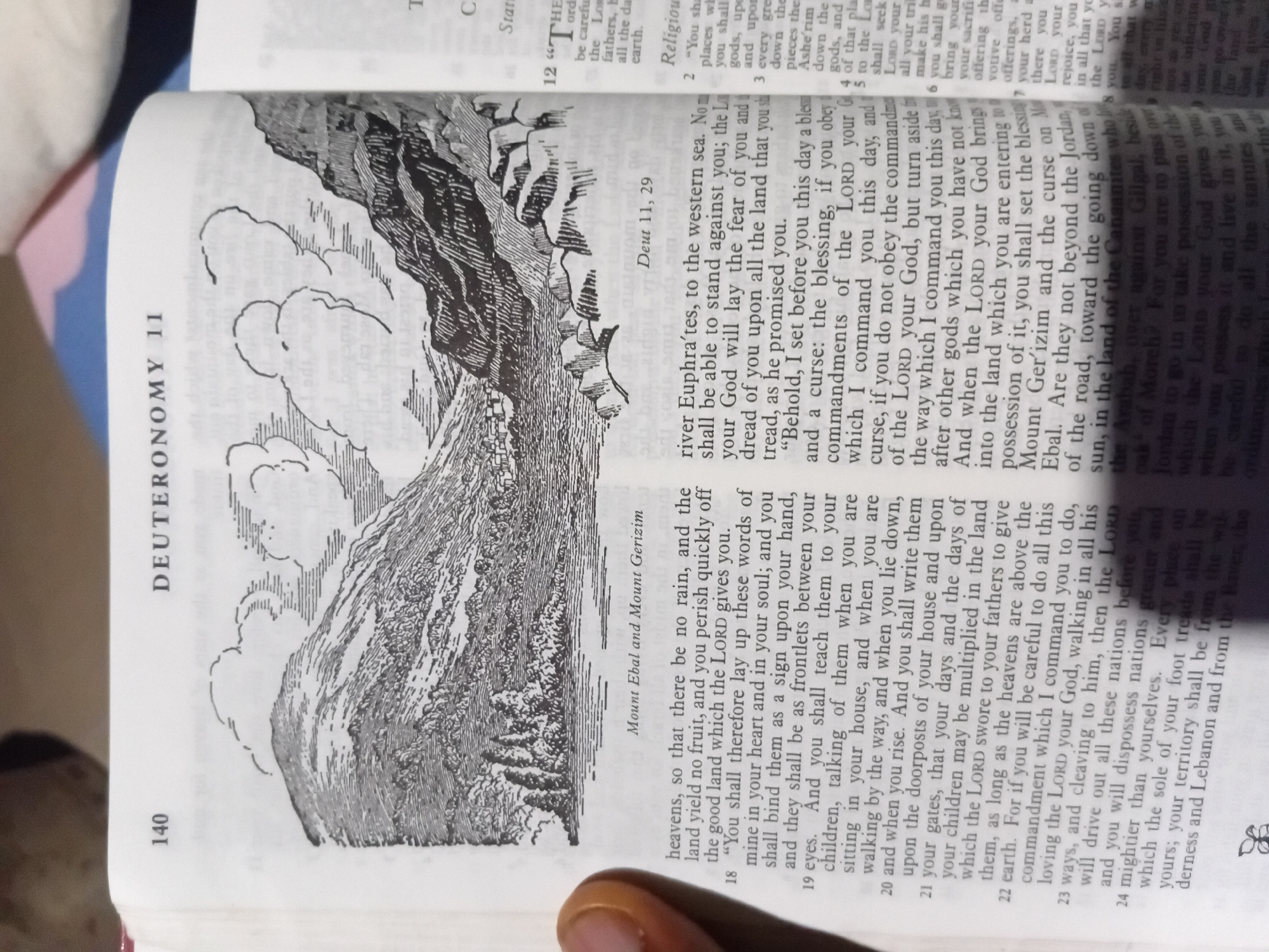 The image shows an open book with text and an illustration. The text is in English and appears to be from the Bible, specifically the book of Deuteronomy. The illustration depicts a mountainous landscape with some structures at the base of the mountains. The page is numbered 140 and titled "DEUTERONOMY 11". The text discusses blessings and curses, obedience to God's commandments, and land promises. The illustration is labeled "Mount Ebal and Mount Gerizim". The bottom right of the image shows more text from what appears to be a commentary or devotional section, with verse numbers like 12 and 2. The image is a close-up shot of the book, with a finger visible on the left edge, suggesting someone is holding the book. The background is blurred, but appears to be indoors, possibly with some patterned fabric visible. There is no indication of the city of Nkpor, Nigeria, in the image or the provided text.
