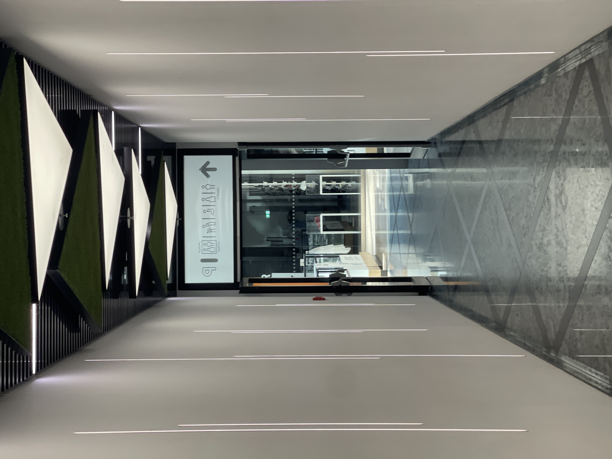 This image depicts a modern, indoor hallway with a unique ceiling design. The hallway is well-lit by a combination of ambient light and vertical LED strips along the walls. The ceiling features a geometric pattern of triangular panels, alternating between white and green, set against a black background with linear lighting. A prominent sign with icons indicating restrooms and an arrow pointing forward is mounted above an entryway. Through the glass doors of the entryway, displays of merchandise and a person can be seen. The floor is reflective, with a pattern of polished stone tiles. The scene appears to be located in a public space, possibly a shopping center or an exhibition hall in Bratislava, Slovakia, given the context of the provided information. There are no individuals clearly visible in the hallway itself, but a figure is present within the space beyond the entryway.