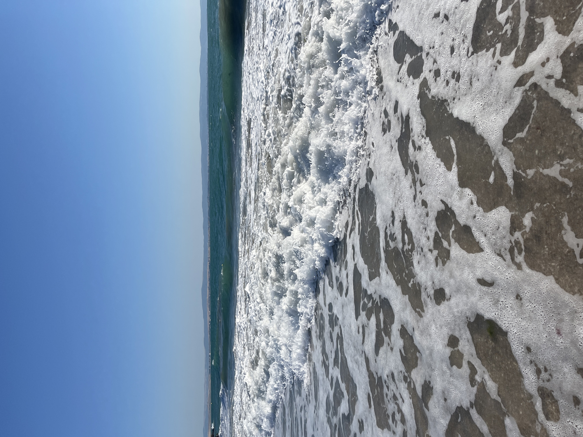 A clear, cloudless sky stretches above a serene beach scene in Pomorie, Bulgaria. The turquoise sea, with waves gently breaking into white foam, dominates the lower half of the image. The shoreline is a mix of sand and shallow water, where the foamy surf creates abstract patterns as it recedes. In the distance, a faint line of hills suggests the coastline of Pomorie. There are no people or man-made objects visible in the frame. The lighting indicates it is likely daytime, with the sun casting a soft glow on the water and sand.