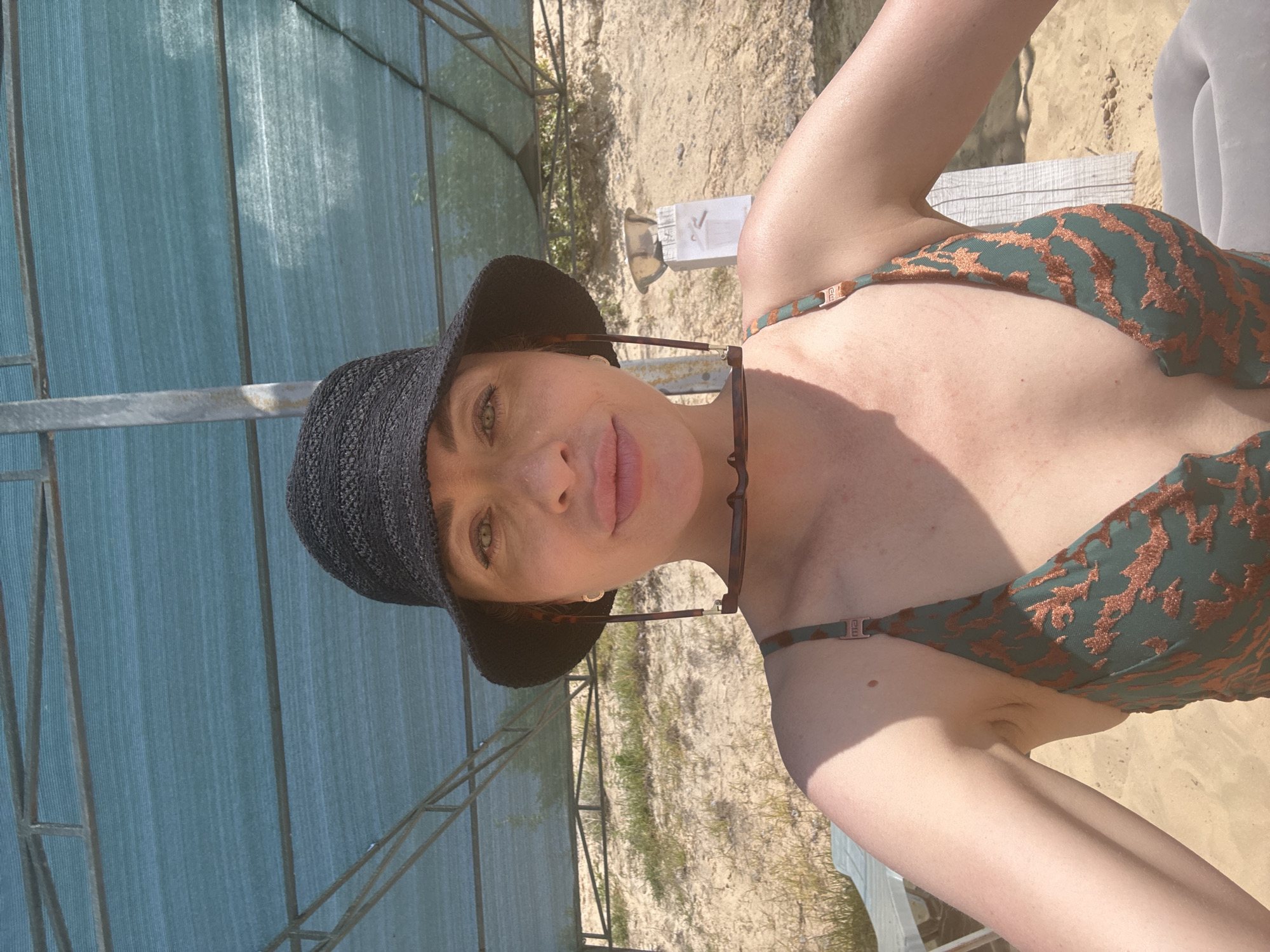 A woman wearing a black bucket hat and a green and brown patterned bikini is pictured outdoors in Yelyzavetivka, Ukraine. She is holding sunglasses by the arms and resting them on the brim of her hat. The woman is looking at the camera with her mouth slightly open. She is positioned in front of a sandy area with some sparse vegetation and a blue tarp overhead supported by metal poles. To her left, a portion of a white lounge chair is visible, and to her right, a white post with a metallic bowl on top can be seen. The scene appears to be a beach or outdoor recreational area.