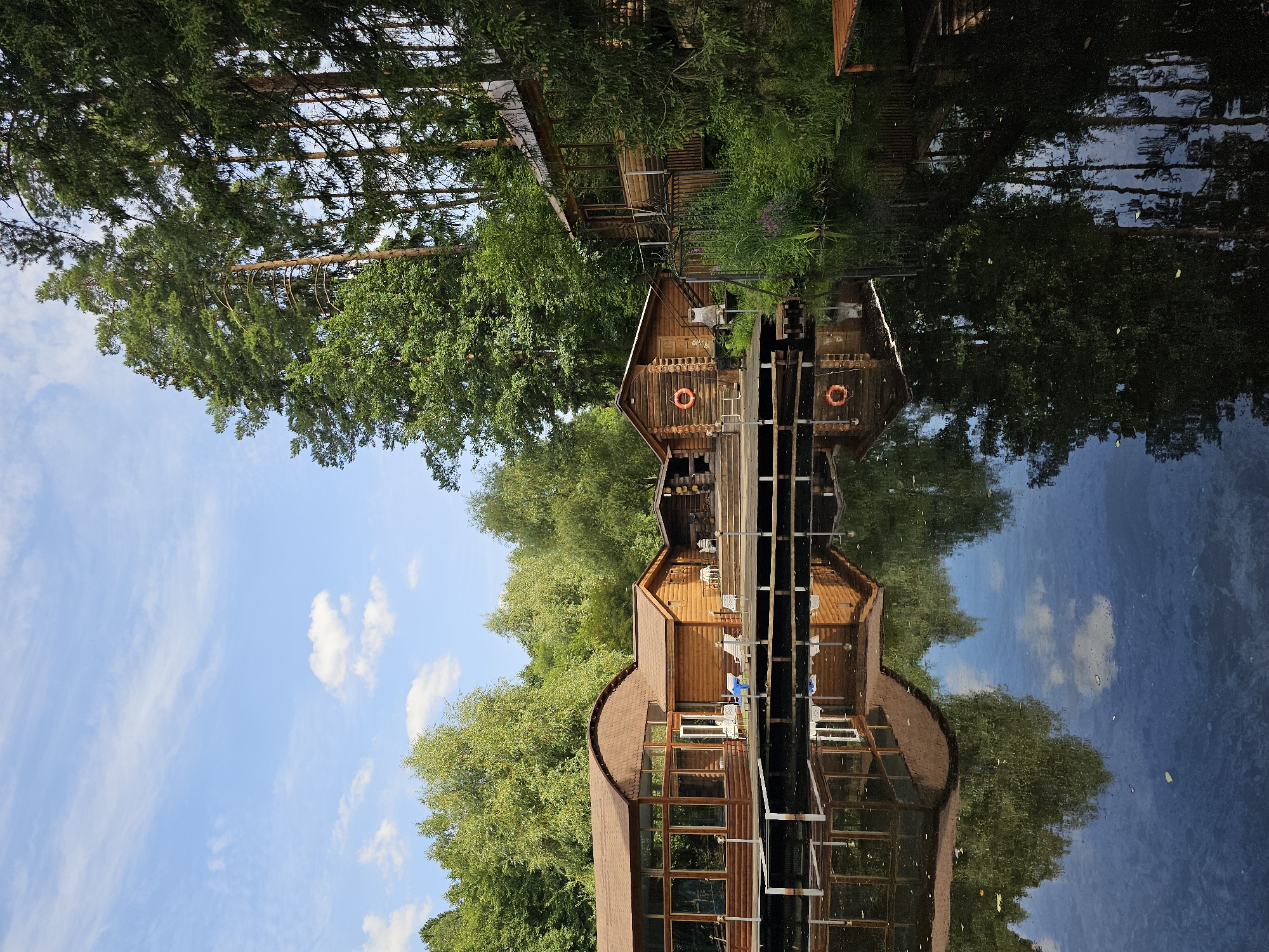 In the city of Ulitkino, Russia, a tranquil waterside scene unfolds. Several wooden buildings, featuring rustic log construction and brown roofs, are situated on a wooden deck extending over a dark body of water. The largest building on the left has a distinctive curved roof and large glass windows. Adjacent to it are smaller log cabins, one of which displays an orange life preserver on its exterior. The buildings are surrounded by lush green trees, with tall pine trees dominating the background on the right. The water perfectly reflects the buildings and the surrounding foliage, creating a mirror image that adds to the serene atmosphere. The sky above is a clear blue, dotted with a few white clouds. The scene appears to be a resort or recreational area, possibly a spa or a lodge, given the presence of lounge chairs and the overall ambiance. There are no people visible in the image.