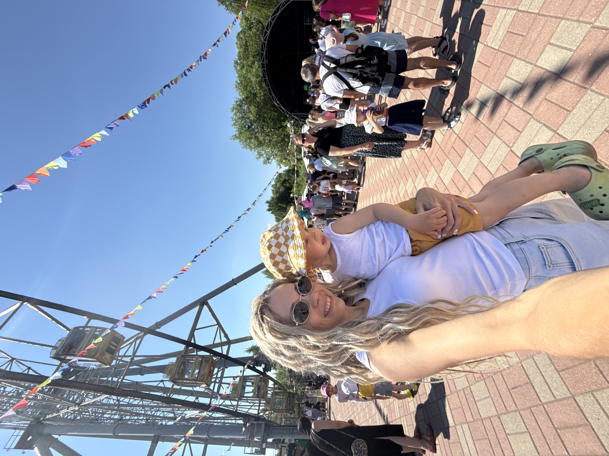 In an outdoor amusement park setting under a clear blue sky, a woman with long blonde dreadlocks and sunglasses holds a young child. The woman is wearing a white tank top and denim shorts, while the child is wearing a white tank top and brown shorts, with a checkered hat. In the background, a Ferris wheel is visible, adorned with colorful triangular flags. A crowd of people are gathered in the park, with a stage set up in the distance. The ground is paved with interlocking bricks in shades of pink and tan. The image appears to be taken on a sunny day.