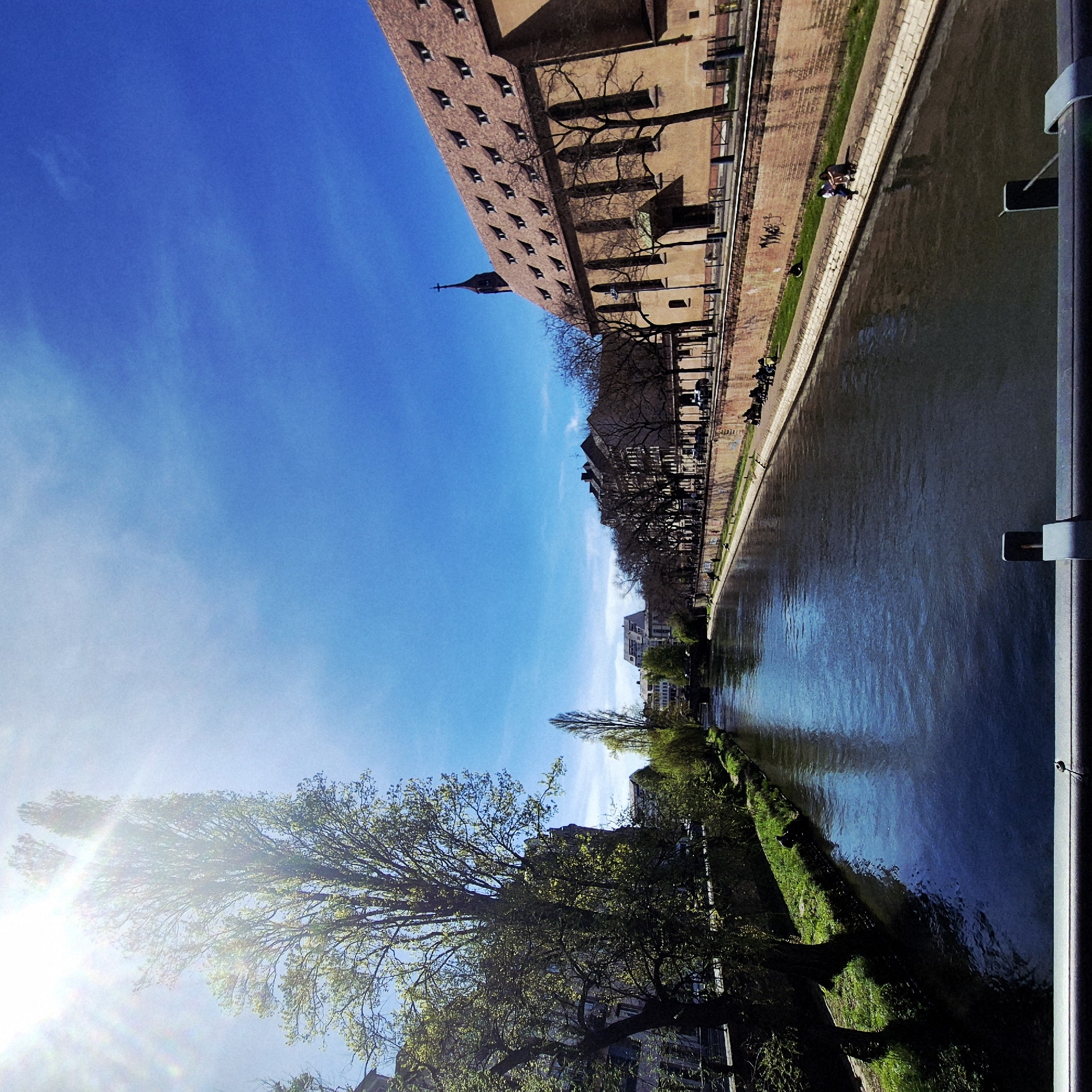 A high-angle, slightly canted perspective depicts a canal in Strasbourg, France, reflecting a clear blue sky. Along the canal's edge, a grassy bank lines a paved walkway where pedestrians stroll. A large brick building with multiple windows stands adjacent to the walkway. Trees with budding leaves are scattered along the canal's edge, with some foliage encroaching on the walkway. The sun glares from the lower-left corner, obscuring details in the trees and buildings. A metal railing is visible on the right side, suggesting the viewpoint is from a bridge or elevated platform. The waterway reflects the sky and surrounding buildings, creating a mirror effect.