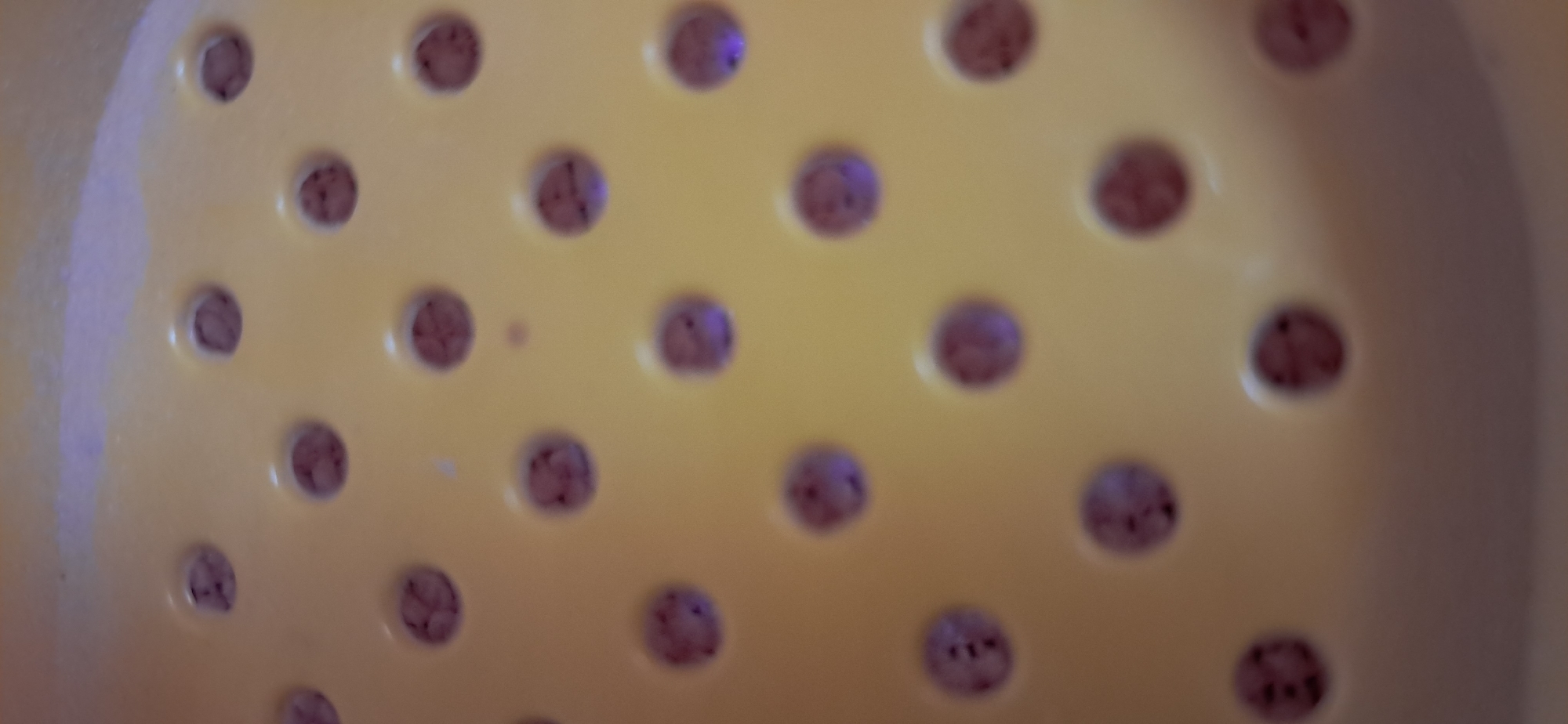 The image shows a close-up, overhead view of a yellow plastic colander with numerous circular holes. Some of the holes appear to contain small, dark specks or debris. The lighting in the image creates highlights and shadows on the textured surface of the colander. The background is out of focus and appears to be a blurred, light-colored surface, possibly a table or countertop. There are no discernible features that would allow for identification of a specific location within the city of Yazy, Turkmenistan. The scene is devoid of any human presence or activity.