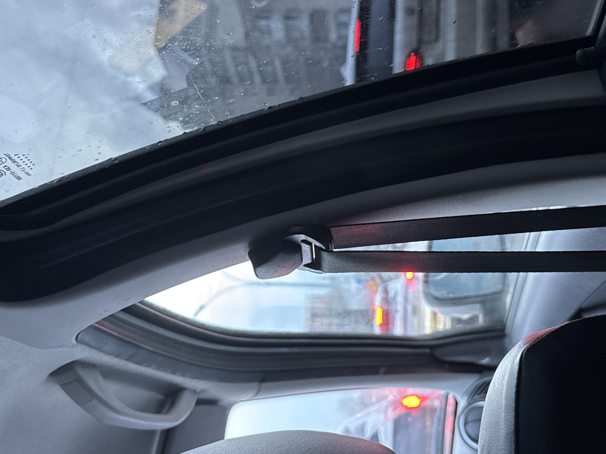 The image offers an interior perspective from a vehicle, looking upwards and forwards through a sunroof. The sunroof's frame is prominent, and its glass is covered in numerous water droplets, indicating rain or recent precipitation.
Inside the car, a light grey ceiling and a matching grab handle on the right side are visible. A black strap system, possibly part of a retractable sunshade mechanism, is attached to the sunroof frame, featuring a central buckle or clip. The top of a car seat, likely dark-colored, can be seen in the lower right corner, partially obscured.
Through the wet sunroof and windshield, the scene outside appears to be an urban environment in Perm, Russia. Multiple red brake lights from other vehicles are visible, suggesting the car is stopped or moving slowly in traffic. Blurred reflections on the glass hint at surrounding buildings or other cars. The lighting is subdued, characteristic of an overcast day.
Text visible on the upper left corner of the sunroof glass, appearing upside down and mirrored due to the reflection, includes "881110-3C0" and the word "TEMPERED." Other faint, unreadable characters and a logo are also present. The overall impression is a moment captured from inside a car during a rainy day in the city traffic.