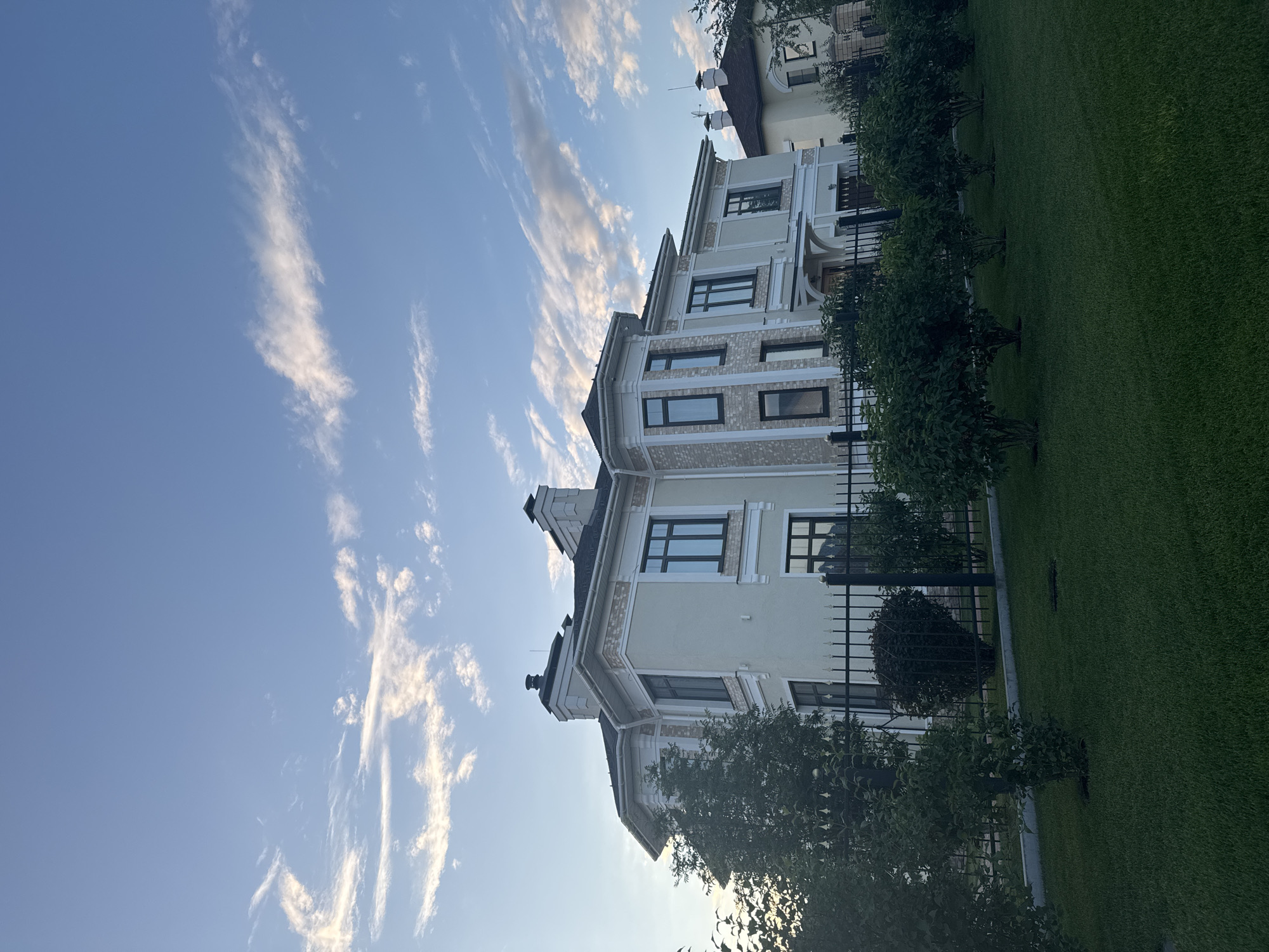 This media file displays a modern, multi-story house with a light-colored facade and decorative stone accents. The architecture features large, dark-framed windows and a steeply pitched roof with intricate detailing. In front of the house, a well-maintained green lawn is bordered by a black metal fence and several neatly trimmed bushes and small trees. The sky above is a clear blue, with wispy, elongated white clouds streaking across it, suggesting a bright, possibly late afternoon or early morning sky. The lighting indicates it is daytime, with the sun casting soft light and shadows. The scene is serene and suggests a residential setting, possibly a suburban or upscale neighborhood. The overall impression is one of order, modernity, and calm. The image appears to be taken in Yekaterinburg, Russia. No people are visible in the frame.