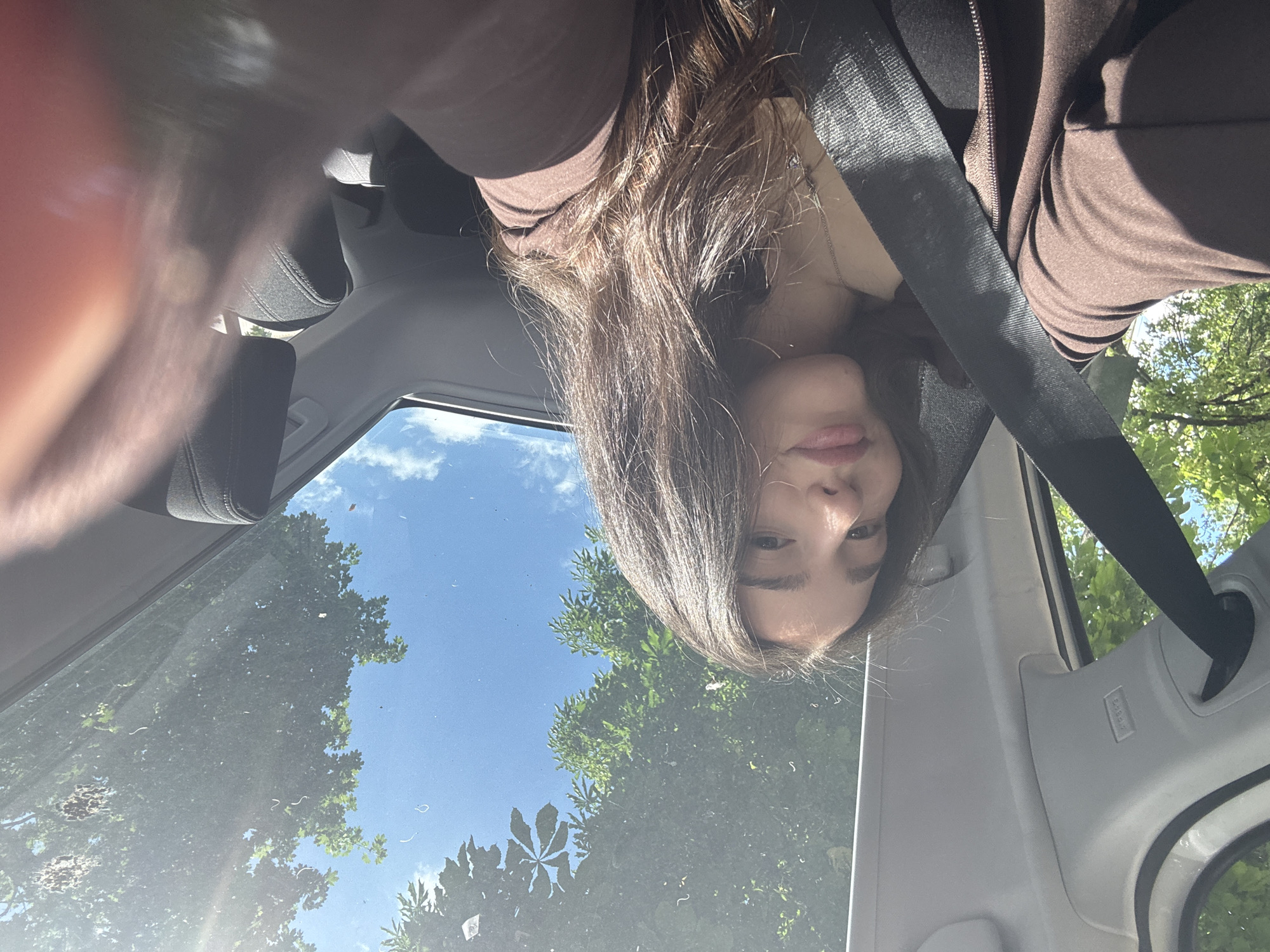A selfie-style image taken inside a car. The subject is a young woman with long brown hair, wearing a brown jacket and a seatbelt. She appears to be sitting in the driver's seat. The car's sunroof is open, revealing a view of trees and a blue sky. The image is taken from a low angle, looking upwards. The city and location are unknown.