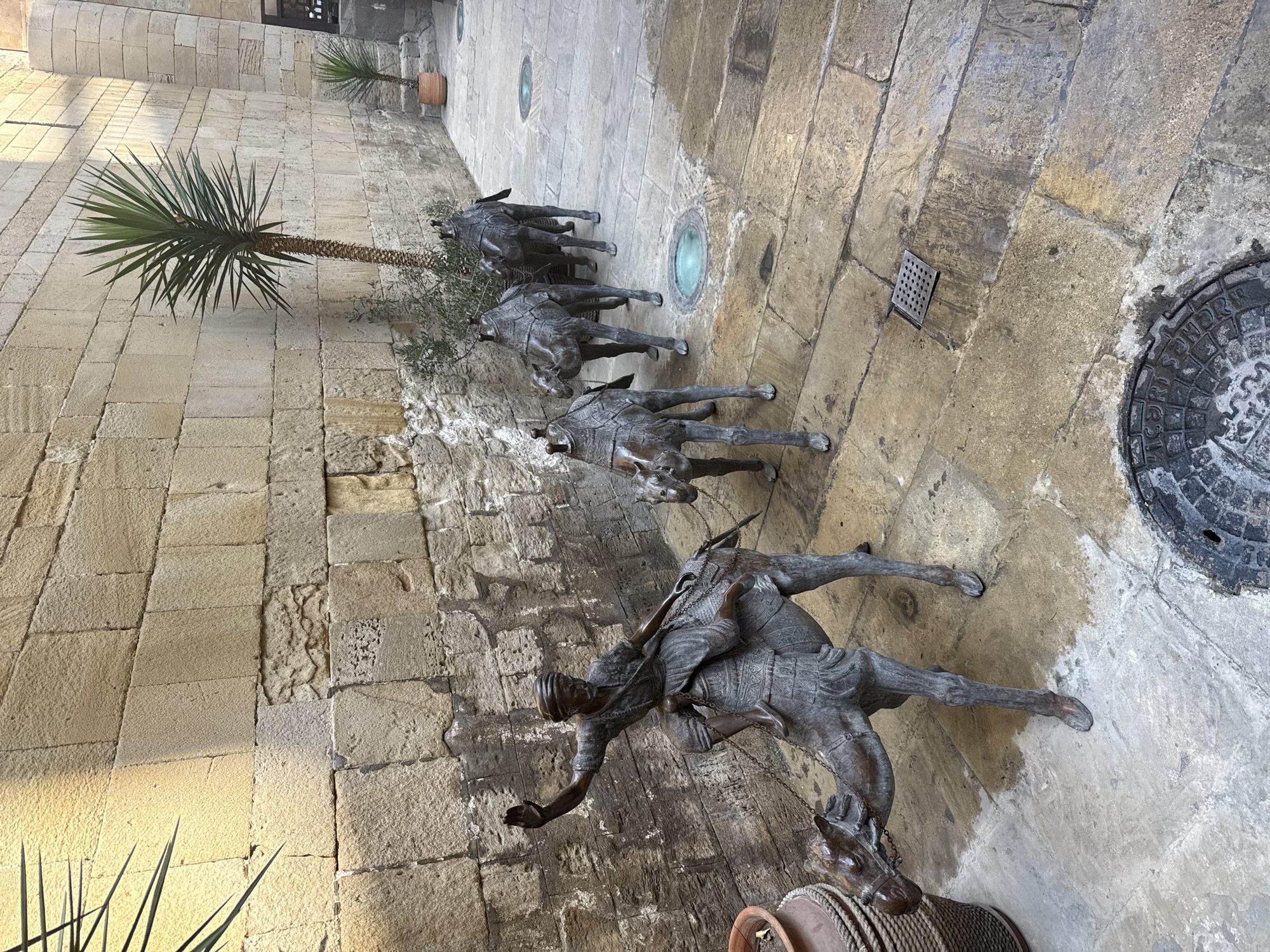 The image depicts a narrow alleyway in Sabail Raion, Azerbaijan, paved with stone slabs. The walls are constructed from rough-hewn stone blocks. The scene is dominated by a line of bronze sculptures of camels, with a rider on the first camel. The rider, wearing traditional attire, is depicted with one hand raised, as if waving. The camels are adorned with ornate saddles and harness. In the background, a tall palm tree stands next to a smaller potted plant, and a second, smaller palm tree is visible further down the alley. The ground is wet, suggesting recent rain or cleaning. Several circular grates or manhole covers are embedded in the pavement. The overall impression is of a historical or cultural site, possibly an art installation within the city's old town.