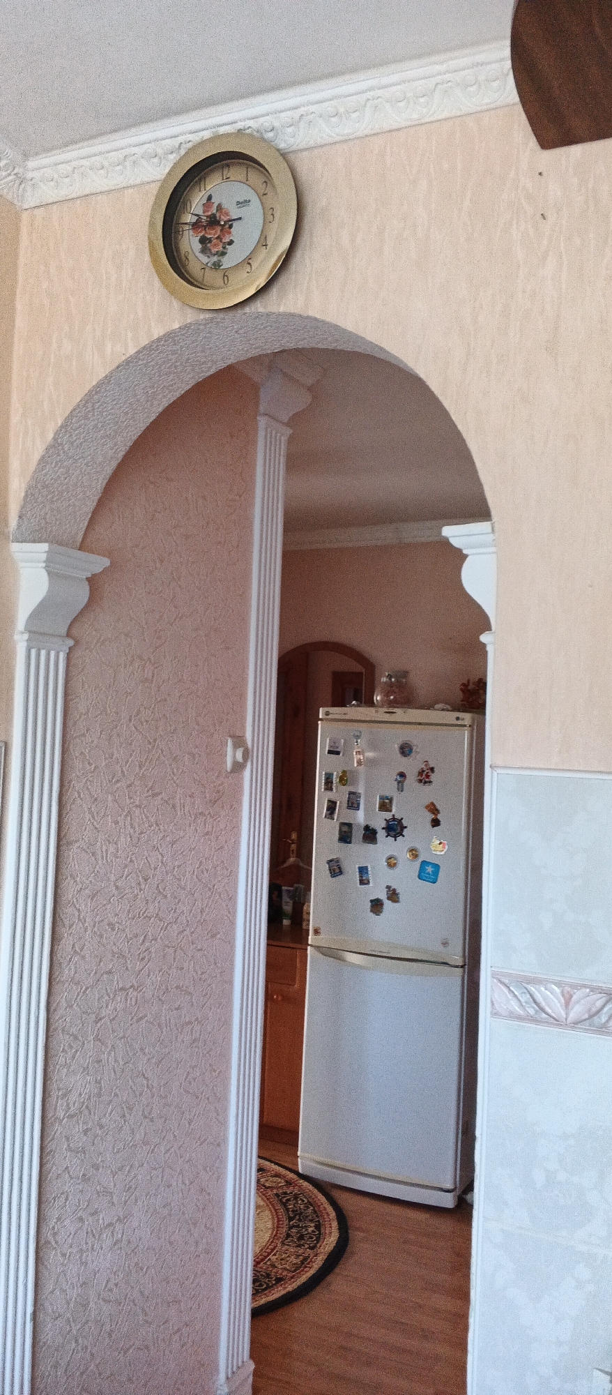 The image shows an interior scene, possibly a home, with a clock on the wall above an archway. The archway frames a doorway that leads into another room where a refrigerator is visible. The refrigerator is adorned with numerous magnets. The overall impression is that of a domestic environment. The provided metadata states the image was taken in Mukachevo, Ukraine.