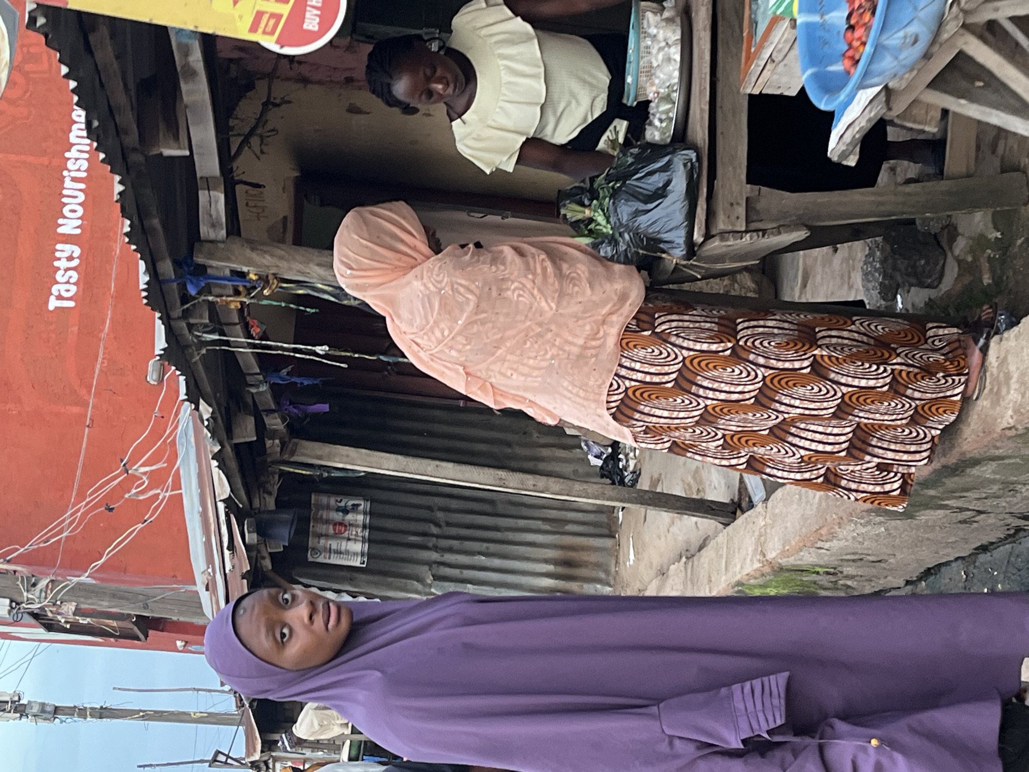 The image captures a street market scene in Jos, Nigeria. In the foreground, a young woman wearing a purple hijab looks directly at the camera with a surprised expression. Behind her, a woman in a peach-colored hijab and patterned skirt is interacting with a vendor. The vendor, a woman with dark skin and a light-colored ruffled top, is behind a stall, holding a bunch of green vegetables wrapped in plastic. The market appears to be made of makeshift structures with corrugated metal roofing and wooden supports. Wires are visible strung across some of the structures, suggesting an informal electrical setup. In the background, a blue basin filled with red fruits and a basket of white items are visible on a shelf. A red sign with white lettering is partially visible on the left side of the image. The overall atmosphere is that of a bustling, everyday market.