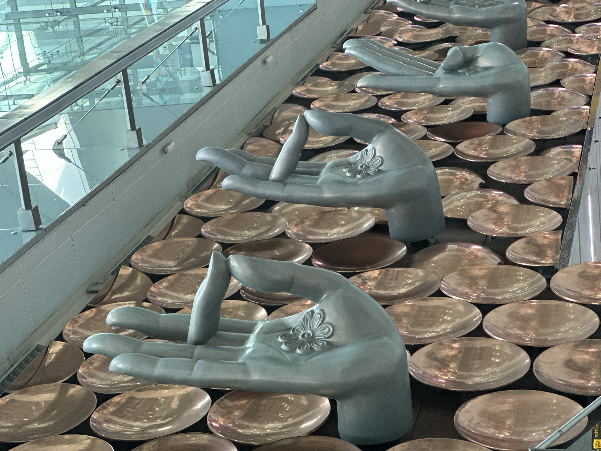 The image captures an art installation located at the Indira Gandhi International Airport in New Delhi, India. The artwork consists of a series of large, silver-colored, stylized hands positioned palm-upwards, each adorned with a floral emblem. These hands are set against a backdrop of numerous small, bronze-colored, reflective circular plates or discs arranged tightly together. The perspective suggests the viewer is positioned above and to the side of the installation. In the background, a modern architectural element is visible, featuring glass panels and metal railings, indicative of an airport terminal setting.