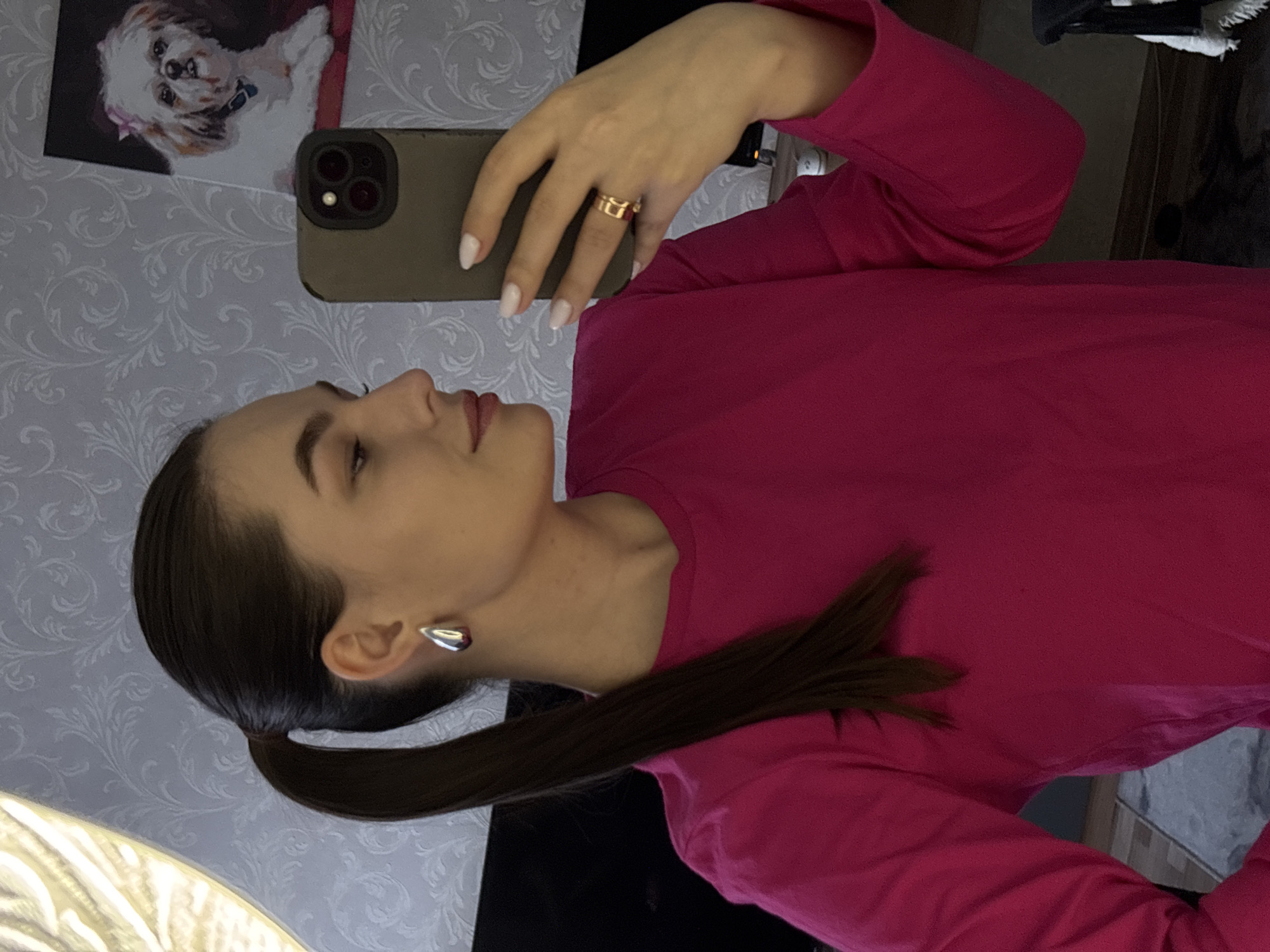 The image displays a young woman, captured in a profile view from her left side. She is holding a dark-cased smartphone with her right hand, positioned as if taking a selfie or viewing its screen. She wears a long-sleeved, magenta-colored top. Her dark hair is neatly pulled back into a low ponytail, and a silver-toned, abstract-shaped earring adorns her left ear. On her right ring finger, a gold-toned ring is visible. Her nails are light-colored, possibly white or pale pink, and almond-shaped. The woman's expression is neutral, with her gaze directed slightly upwards and to the side.
The background features a light-colored wall covered with patterned wallpaper, displaying a subtle damask or floral design. Above where the phone is held, a framed picture of a small white dog, wearing a pink bow and a blue collar, hangs on the wall. The setting is indoors and appears to be well-lit, suggesting either daytime or bright artificial lighting. No text is visible in the image. The scene is set in Chaikovsky, Russia.