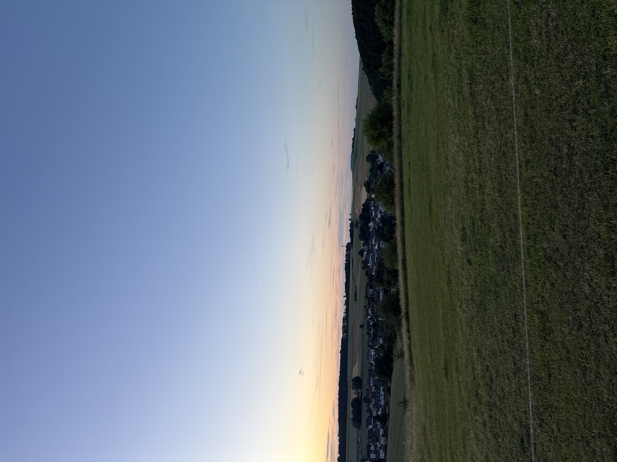 This is a wide-angle, eye-level, exterior shot of a rural landscape near Oberquembach, Germany, captured at dusk. The sky dominates the upper half of the frame, transitioning from a pale blue at the top to soft orange and yellow hues near the horizon. A thin line of clouds stretches across the sky. In the middle ground, a village with houses and red-tiled roofs is nestled among rolling hills. Fields of varying colors, likely farmland, surround the village, with a dark line of trees forming a backdrop. The foreground consists of a grassy field, appearing somewhat unkempt, with a thin wire running horizontally across it. The lighting suggests it is either sunrise or sunset, with the sun low on the horizon. There are no people or animals visible in the image. The overall impression is one of tranquility and natural beauty.