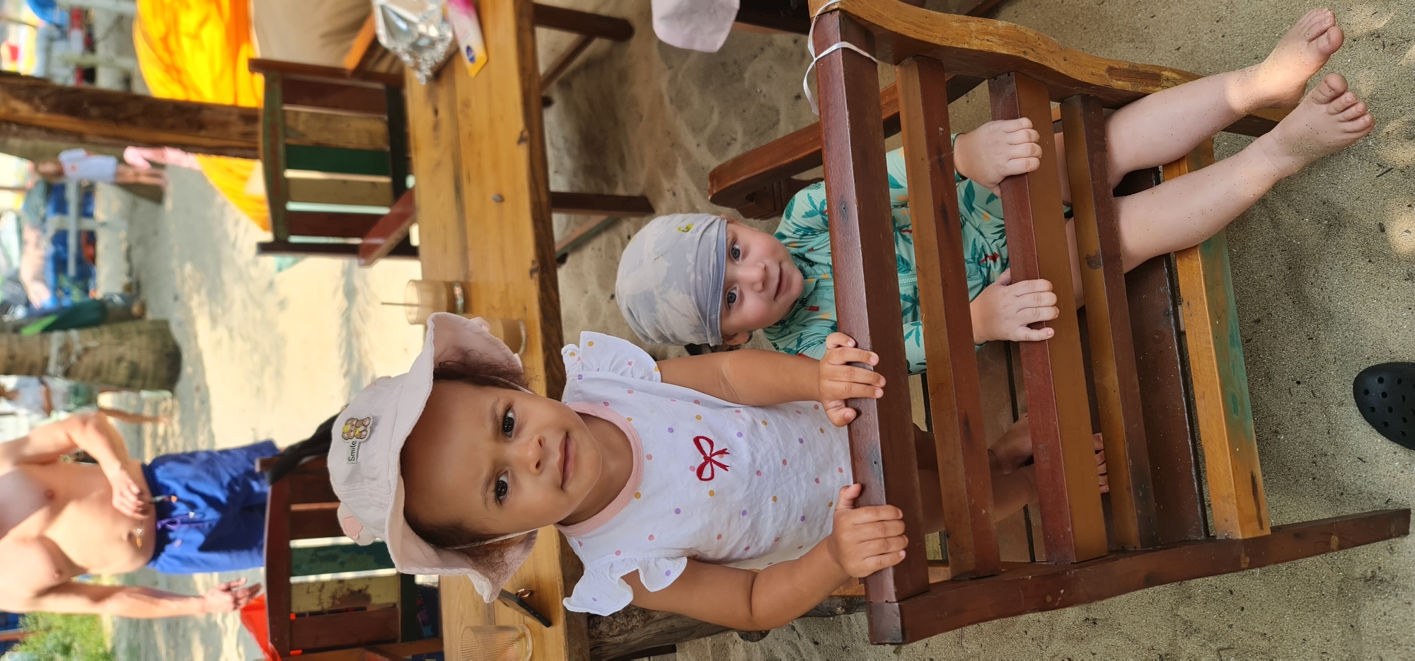 The image depicts two young children at what appears to be a beachside location, possibly in Đà Nẵng, Vietnam, given the sandy environment and tropical vibe. A toddler wearing a white sun hat and a white shirt with colorful polka dots and a red bow is positioned in front, looking directly at the camera. Behind her, another child wearing a light blue patterned head covering and a light green patterned shirt is peering through the slats of a wooden chair. The second child's legs are extended through the slats of the chair. Both children appear to be playing around the chair. The scene includes parts of a wooden table, chairs, and a man in blue swimming trunks standing nearby. The ground is covered with sand. A black shoe is on the right side of the image.