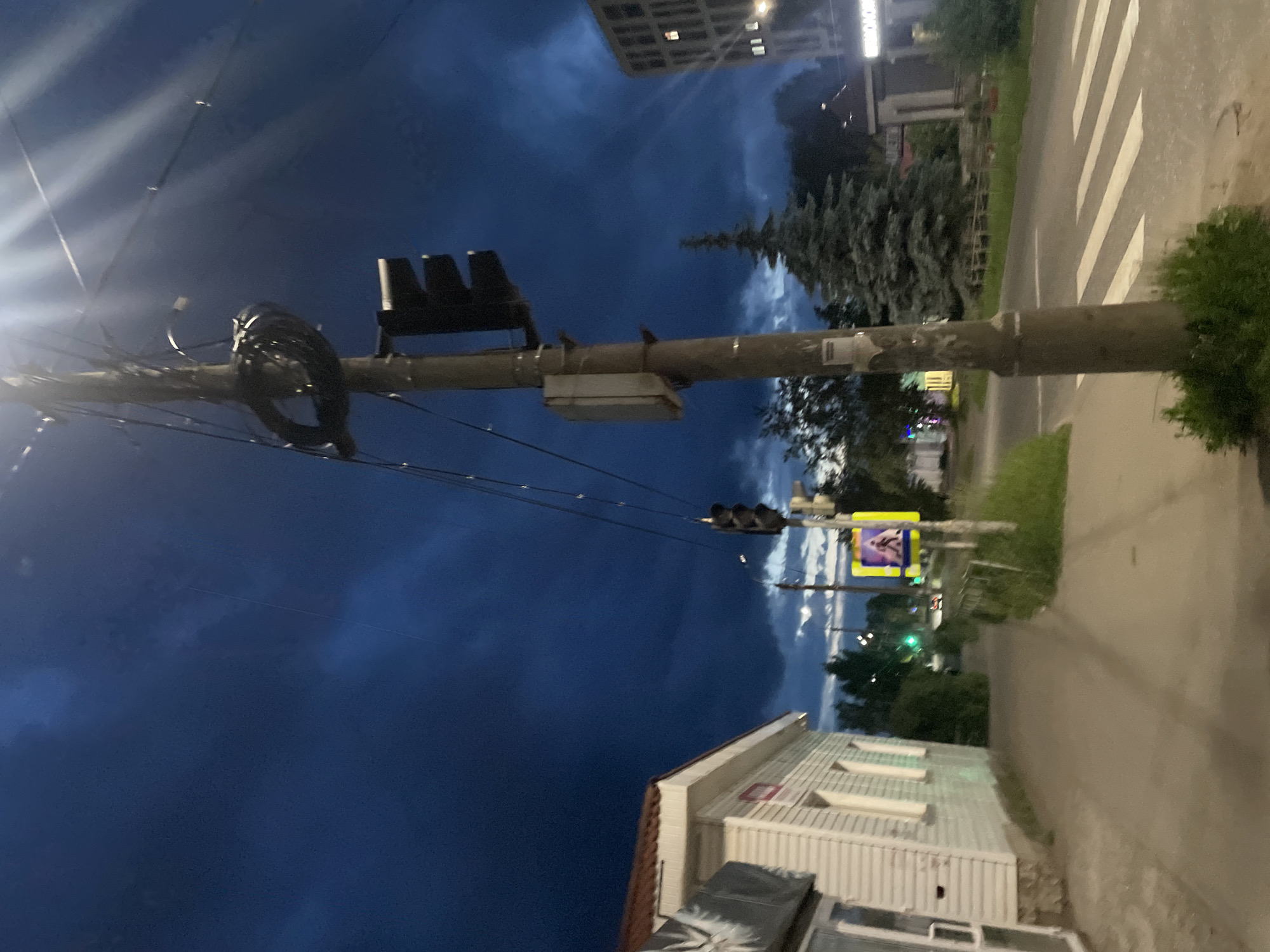The image captures a street scene at dusk or early night in Votkinsk, Russia. The sky is a deep, dark blue, with some lighter patches suggesting clouds. A prominent subject is a utility pole with numerous wires. Attached to this pole are traffic lights, one set of which is visible in the upper left, appearing dark and likely not illuminated. Further down the pole, another set of traffic lights is visible, with a yellow traffic light illuminated, suggesting it is currently active.
To the right of the pole, a pedestrian crossing with white stripes is visible on a paved road. Beyond the road, there's a building with a light-colored, corrugated metal facade and several rectangular skylights. In the background, a multi-story building with visible windows can be seen. A tall, dark green coniferous tree stands between the utility pole and the pedestrian crossing, with other foliage present.
The overall atmosphere is calm and quiet, typical of a street scene after sunset. There are no people clearly visible in the image. The lighting suggests it is twilight or early evening, with artificial lights beginning to appear. The image is oriented vertically, with the pole running from the bottom left towards the upper right. The specific location cues are the Votkinsk, Russia context provided.