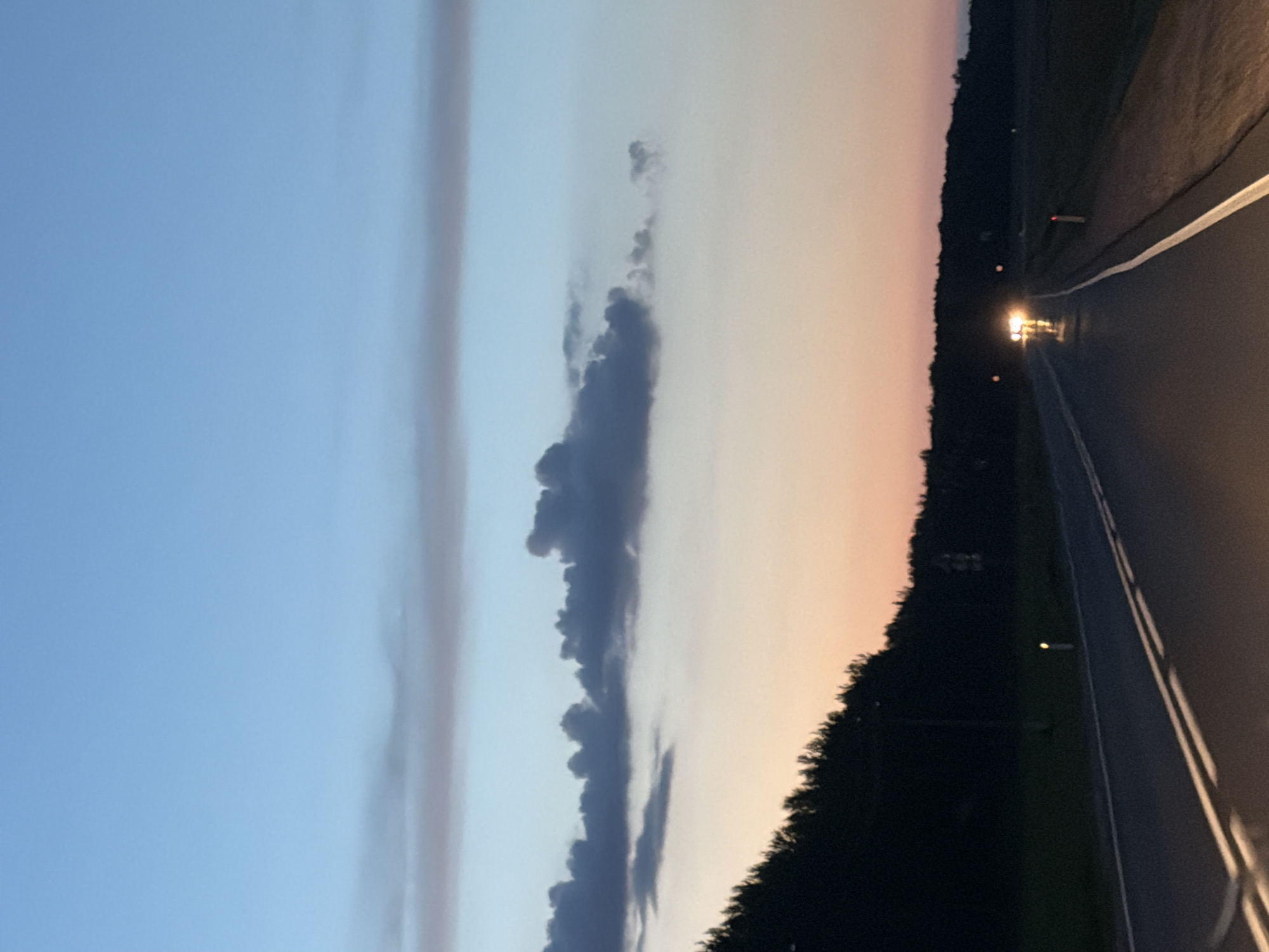 The image depicts a twilight scene on a road in Laishevo, Russia. The sky transitions from a deep blue at the top to hues of orange and pink near the horizon, with scattered clouds. A dark line of trees borders the right side of the road, and fields are visible beyond. The asphalt road, marked with white lines, stretches into the distance. Headlights from an oncoming vehicle illuminate the wet road surface, suggesting recent rain. Several road markers, some with reflective elements, are visible along the side of the road. The overall atmosphere is serene and slightly melancholic, characteristic of the transition from day to night.