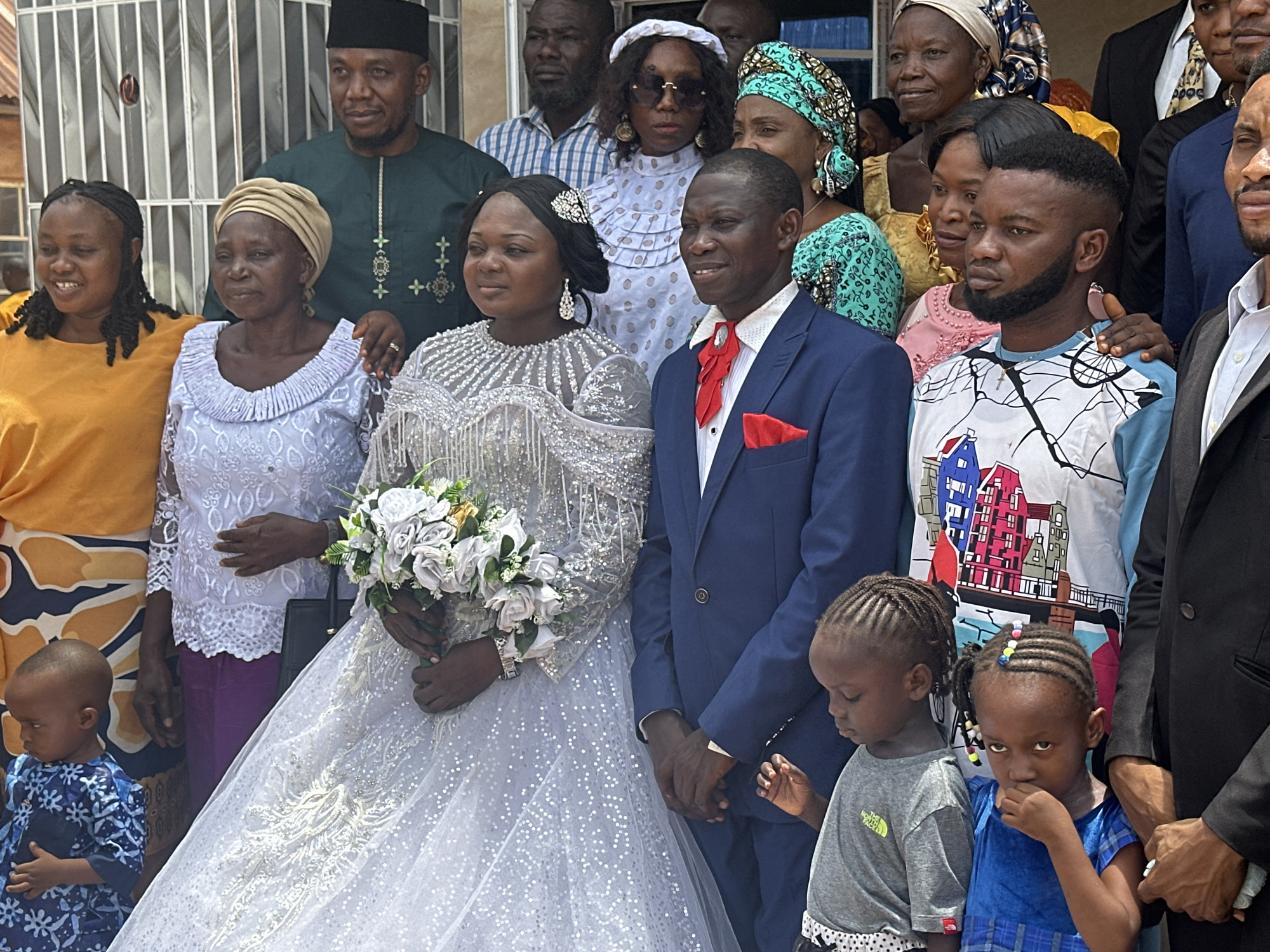 A group of people poses for a photo in Unknown, Unknown. The focal point is a bride in a full white wedding gown with a glittery bodice and a groom in a blue suit with a red bow tie and pocket square. The bride holds a bouquet of white roses. They are surrounded by wedding guests of varying ages and attire, including some in traditional African clothing. Two young children stand at the front of the group. The setting appears to be outdoors, with a building visible in the background.