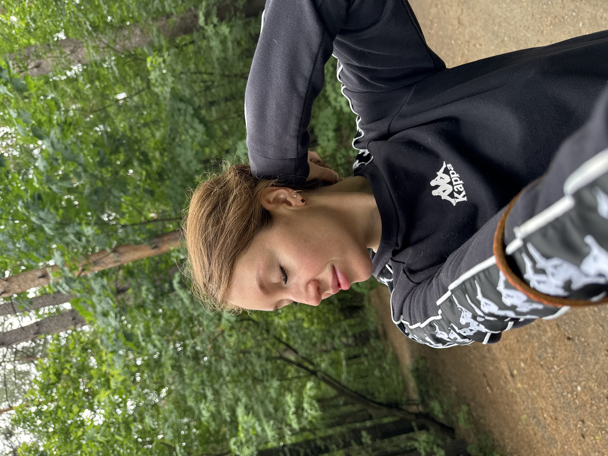 The image shows a young woman with reddish-brown hair, wearing a black Kappa sweatshirt with white detailing on the sleeves. She has her eyes closed and her right hand is touching her head. She is standing in a wooded area with trees and a dirt path visible in the background. The lighting suggests it is daytime. The location is identified as Chaikovsky, Russia.